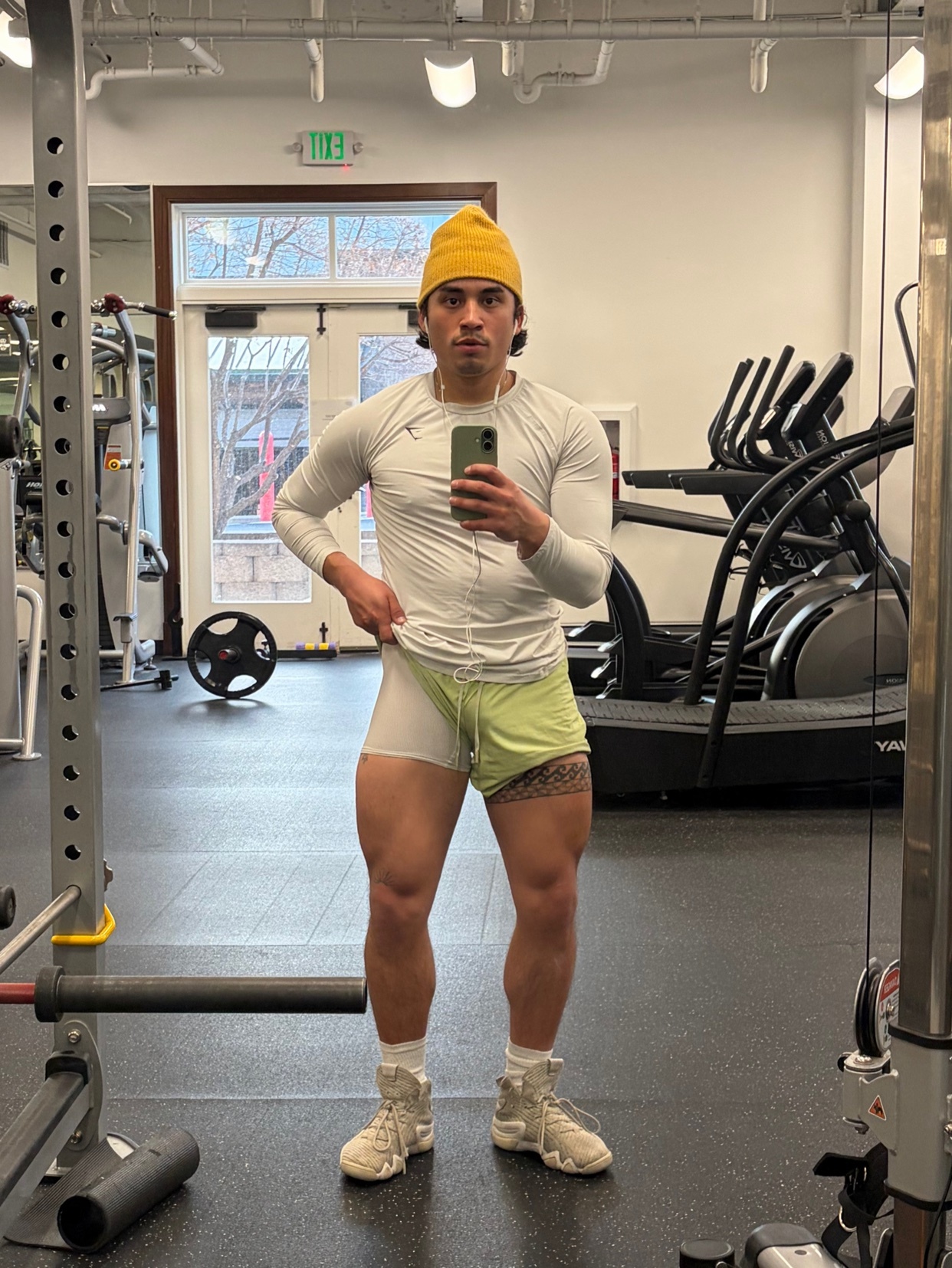 A man in a yellow beanie and a long-sleeved shirt poses for a selfie in a gym. He is wearing light green shorts and standing in front of gym equipment.