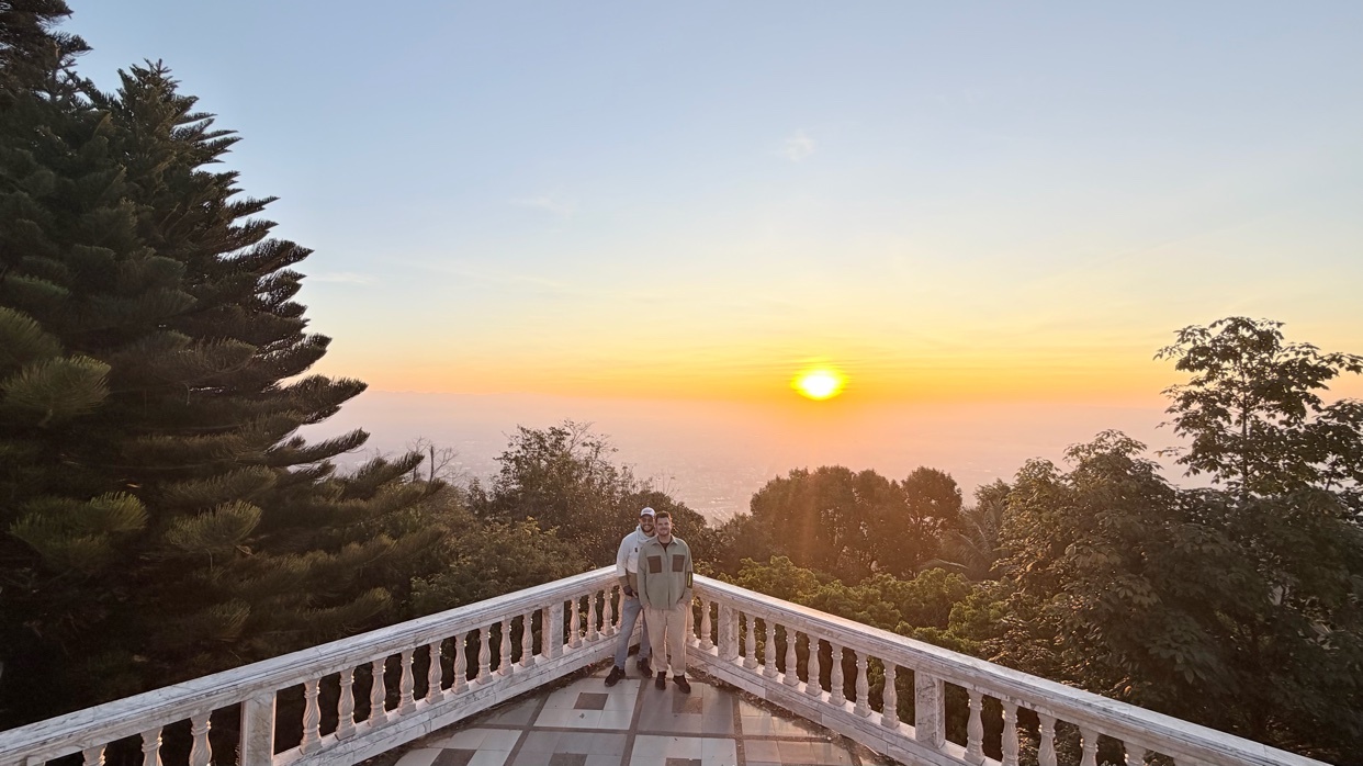 A couple stands on a balcony overlooking a scenic sunset with trees and mountains in the background.