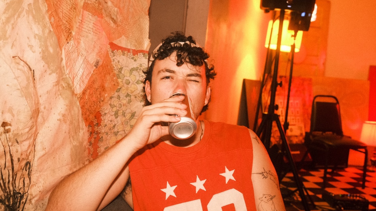 A man with curly hair and a bandana is drinking from a can and winking at the camera. He is wearing a sleeveless orange shirt with white stars and the number '79'.
