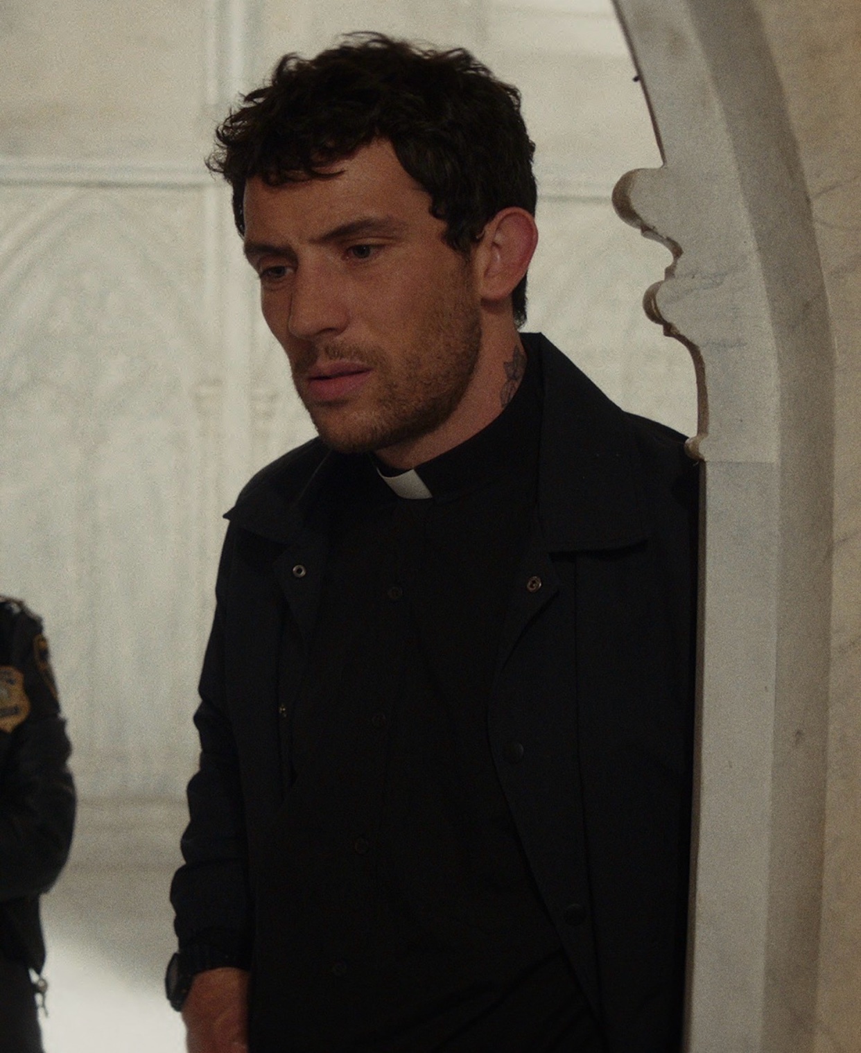 A man in a priest's collar and black suit. He is standing in a doorway.