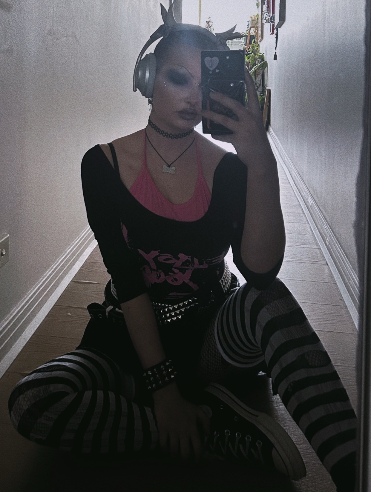 A non-binary person takes a selfie in a hallway. They are wearing a punk-inspired outfit with striped leggings and headphones.