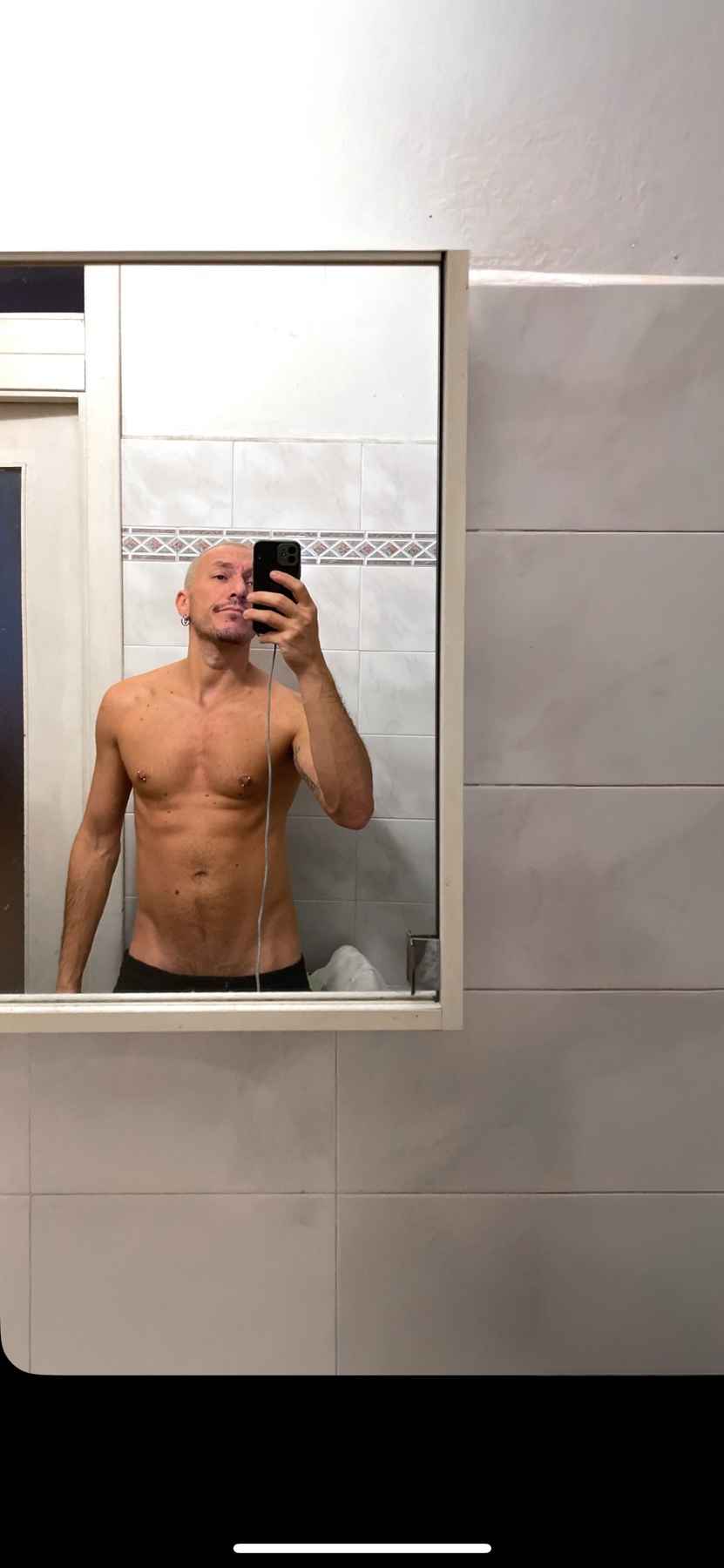 A bald man takes a shirtless mirror selfie in a bathroom. He is flexing his muscular physique.