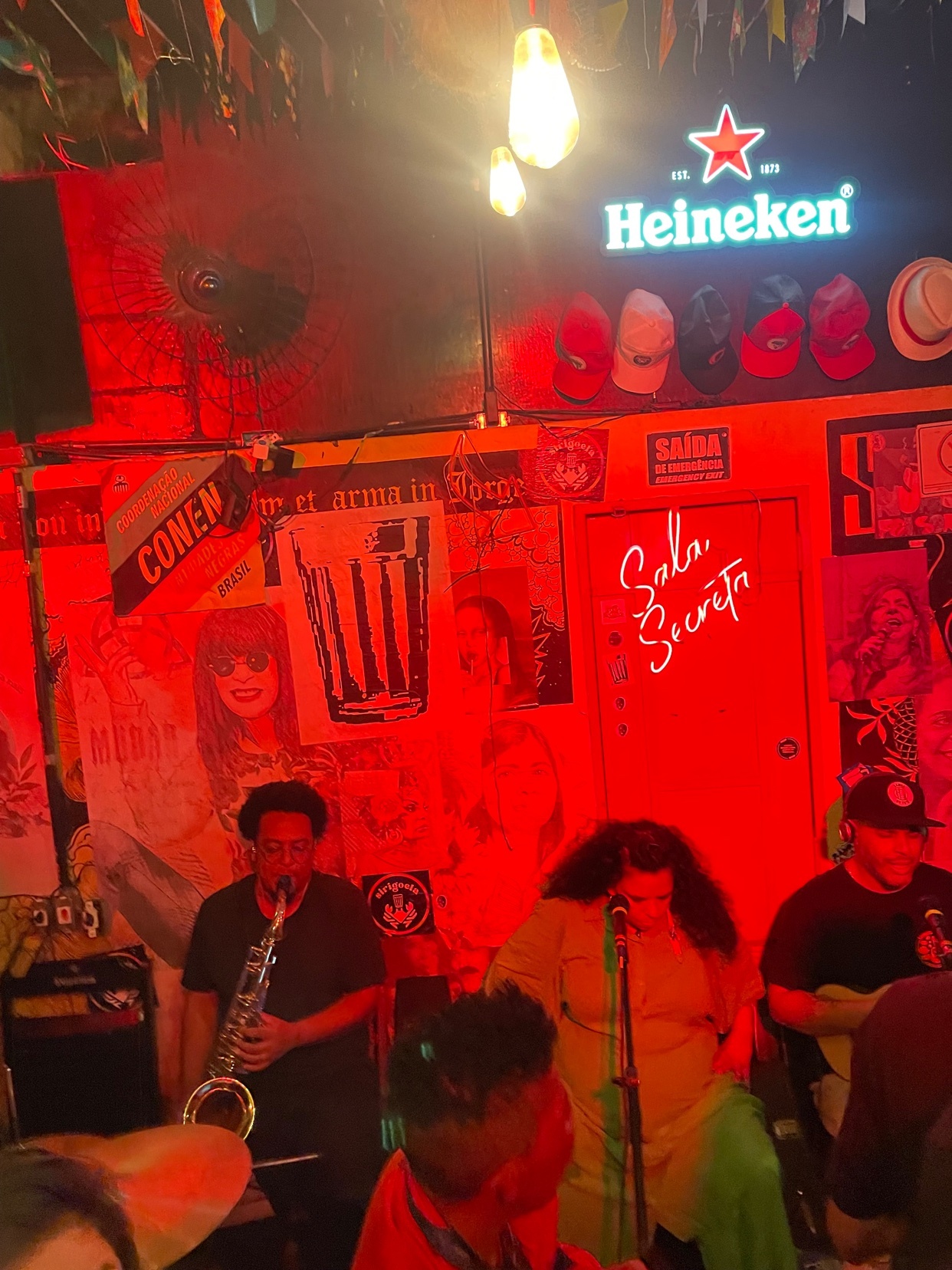 A band performs on a stage in a dimly lit venue. The scene is bathed in red light, with posters and a Heineken sign visible in the background.