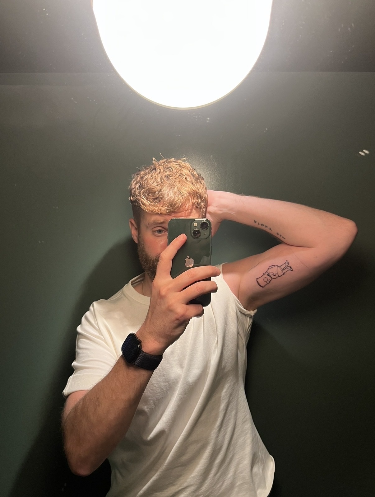A man with blond, curly hair takes a selfie in a mirror, showing off his arm tattoo.