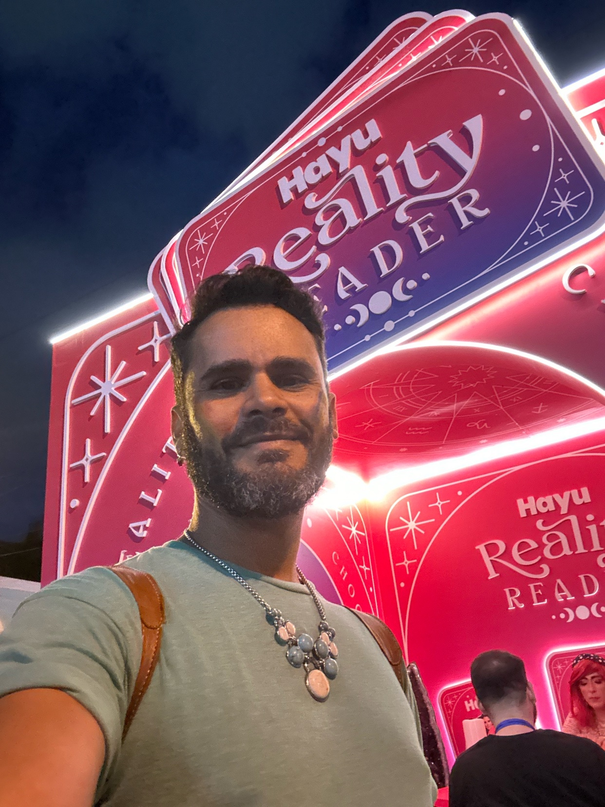 A man with a beard smiles for a selfie in front of a brightly lit sign that reads "Hayu Reality Reader" at night.
