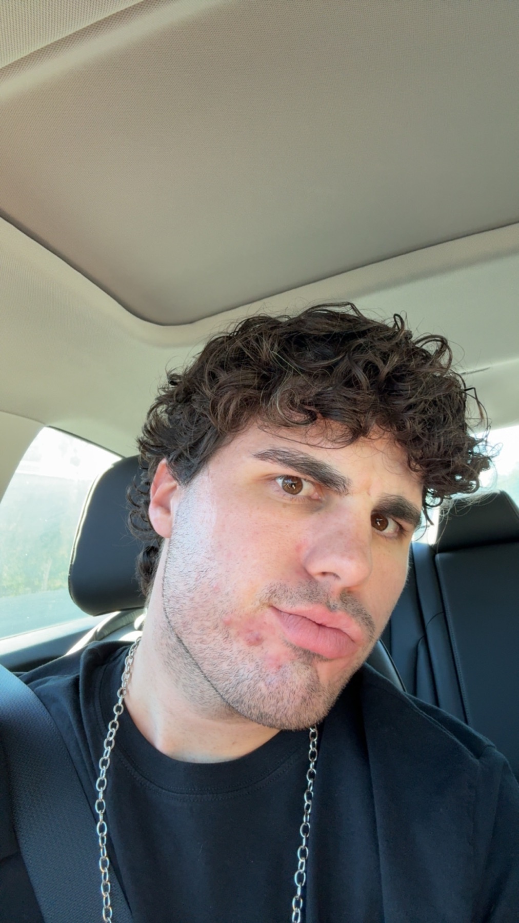 A man with curly hair takes a selfie in a car. He is shirtless and wearing a chain.