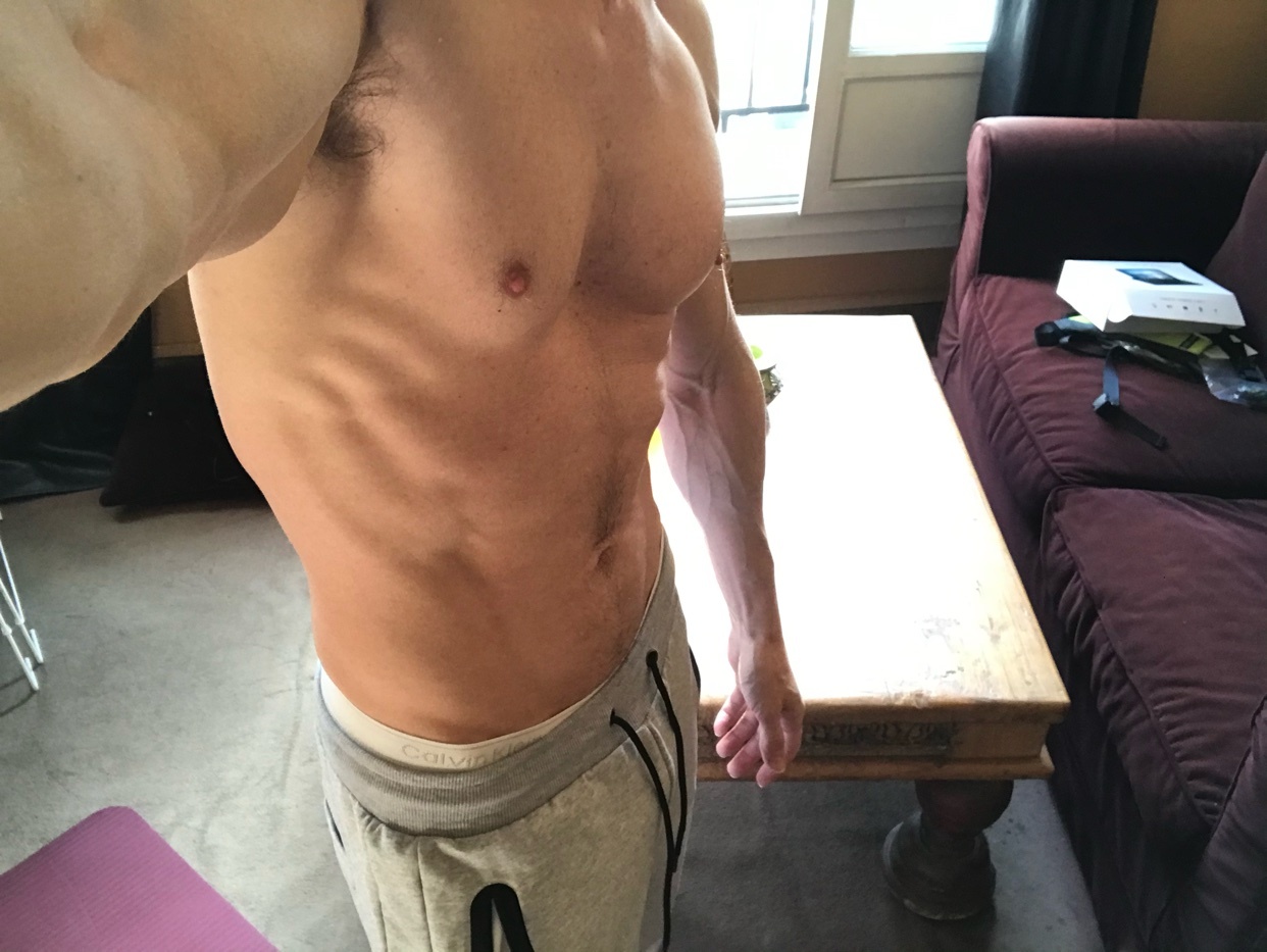 A man shows off his muscular physique in a selfie, highlighting his abs and toned arms. He is wearing grey sweatpants with a white waistband visible.