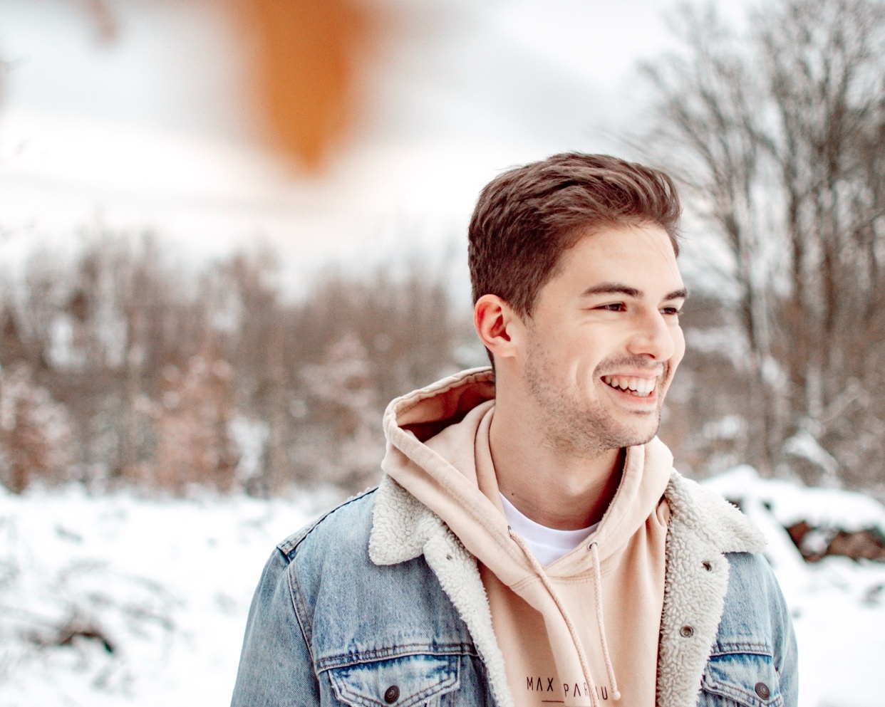 A man smiles in a snowy outdoor setting. He is wearing a denim jacket and a hoodie.