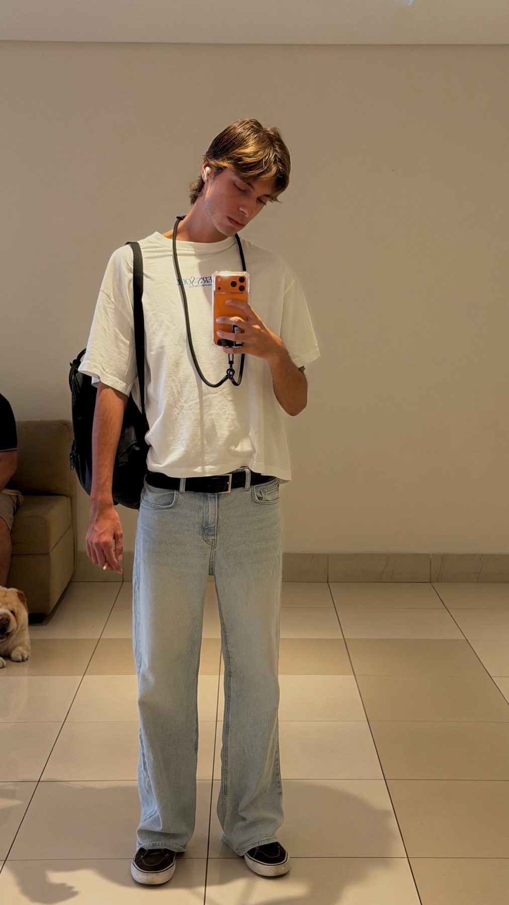 A young man in a white t-shirt and light-wash jeans poses for a selfie in a hallway. He wears a black backpack and earbuds, and holds his phone in his right hand.