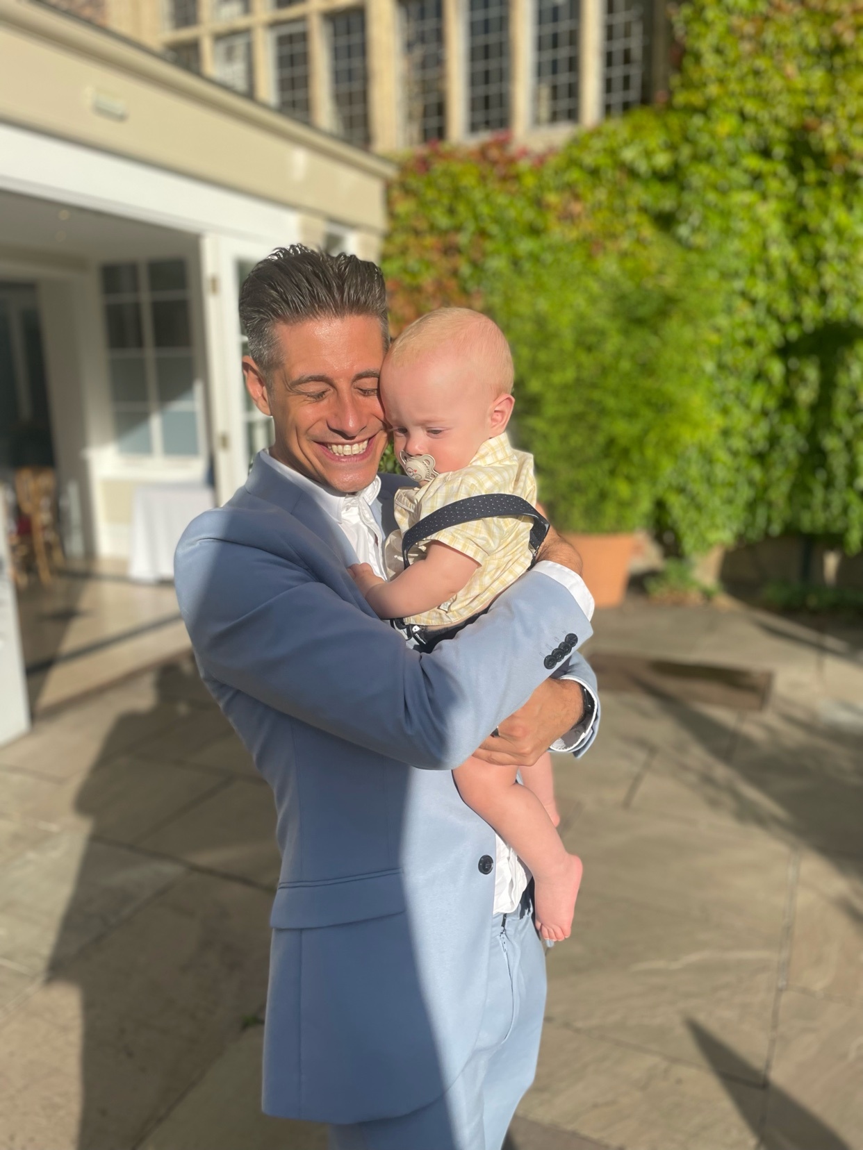 A man in a blue suit holds a baby. They are both smiling in front of a building.