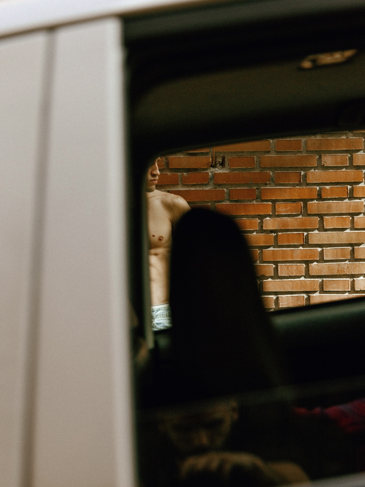 A man stands shirtless in front of a brick wall, viewed through a car window. The image captures a moment of casual intimacy.