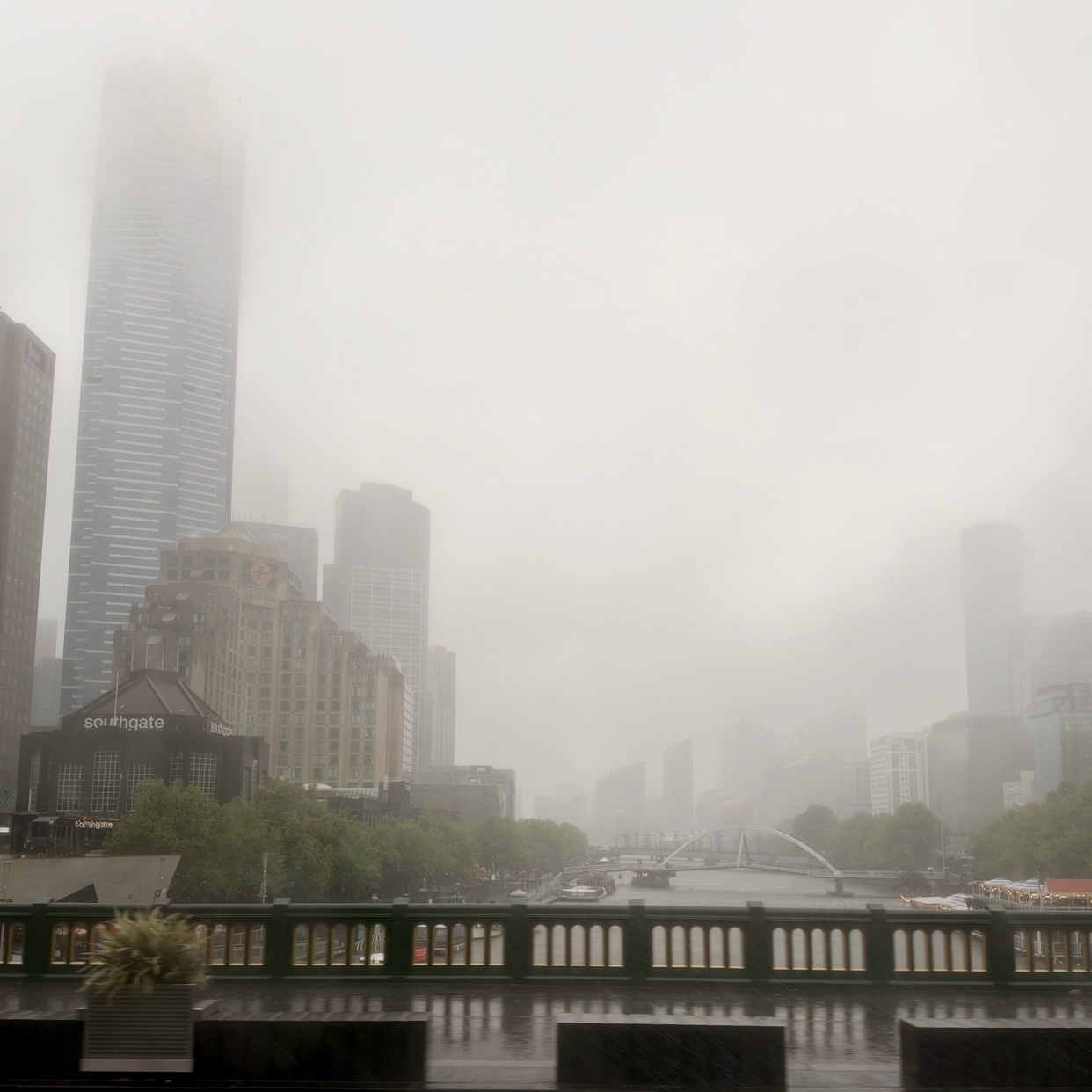 A foggy, overcast day in Melbourne, Australia, with skyscrapers and a bridge visible through the mist. Rain appears to be falling, as indicated by the caption and the wet appearance of the foreground railing.