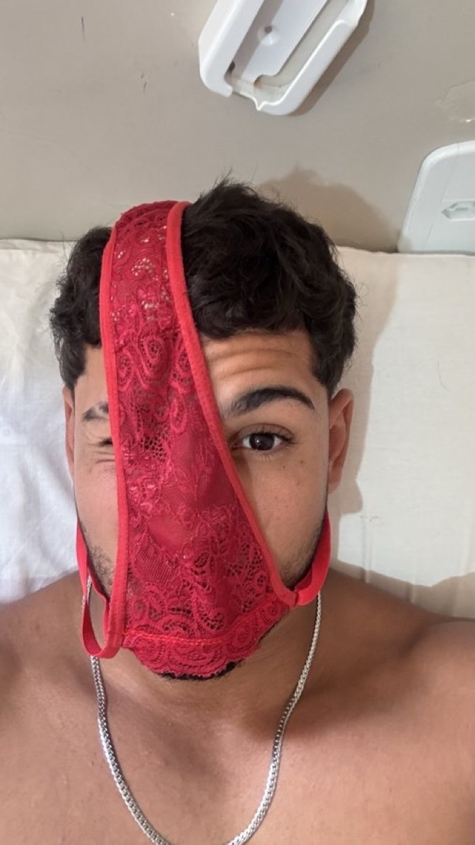 A man covers his face with a red lace thong, leaving one eye visible. He wears a silver chain necklace.