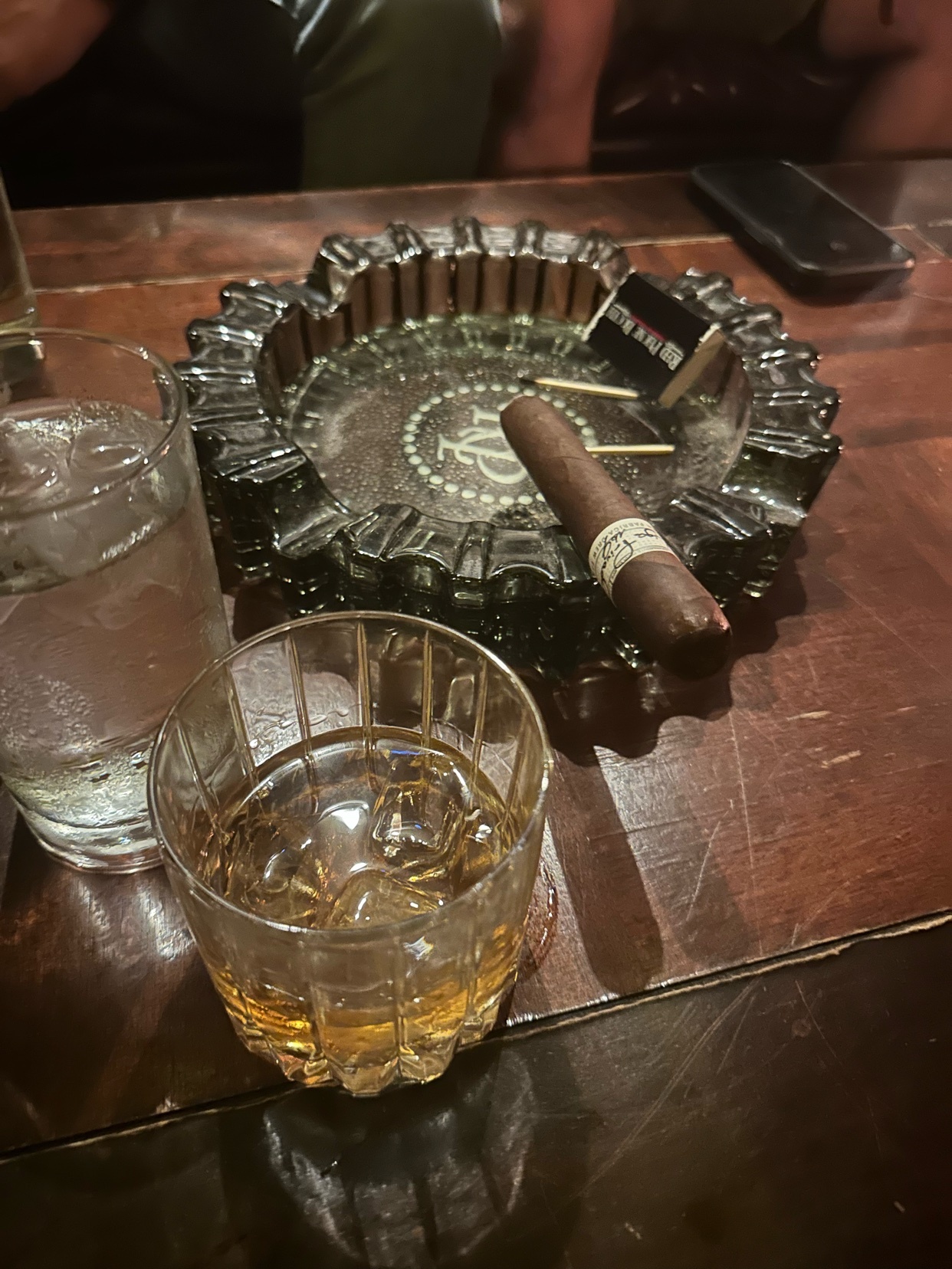 A cigar rests in an ashtray next to a glass of whiskey. The photo captures a speakeasy vibe with a dark, vintage aesthetic.