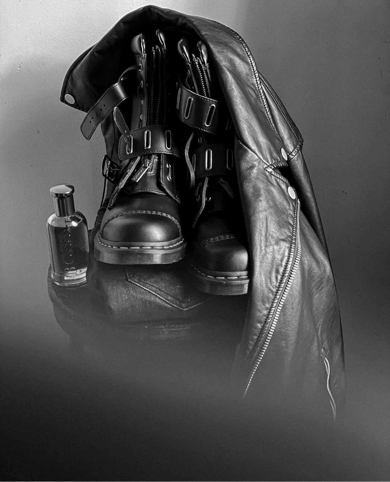 A black and white photo of a pair of boots, a leather jacket, and a bottle of perfume. The items are arranged in a stylish composition.