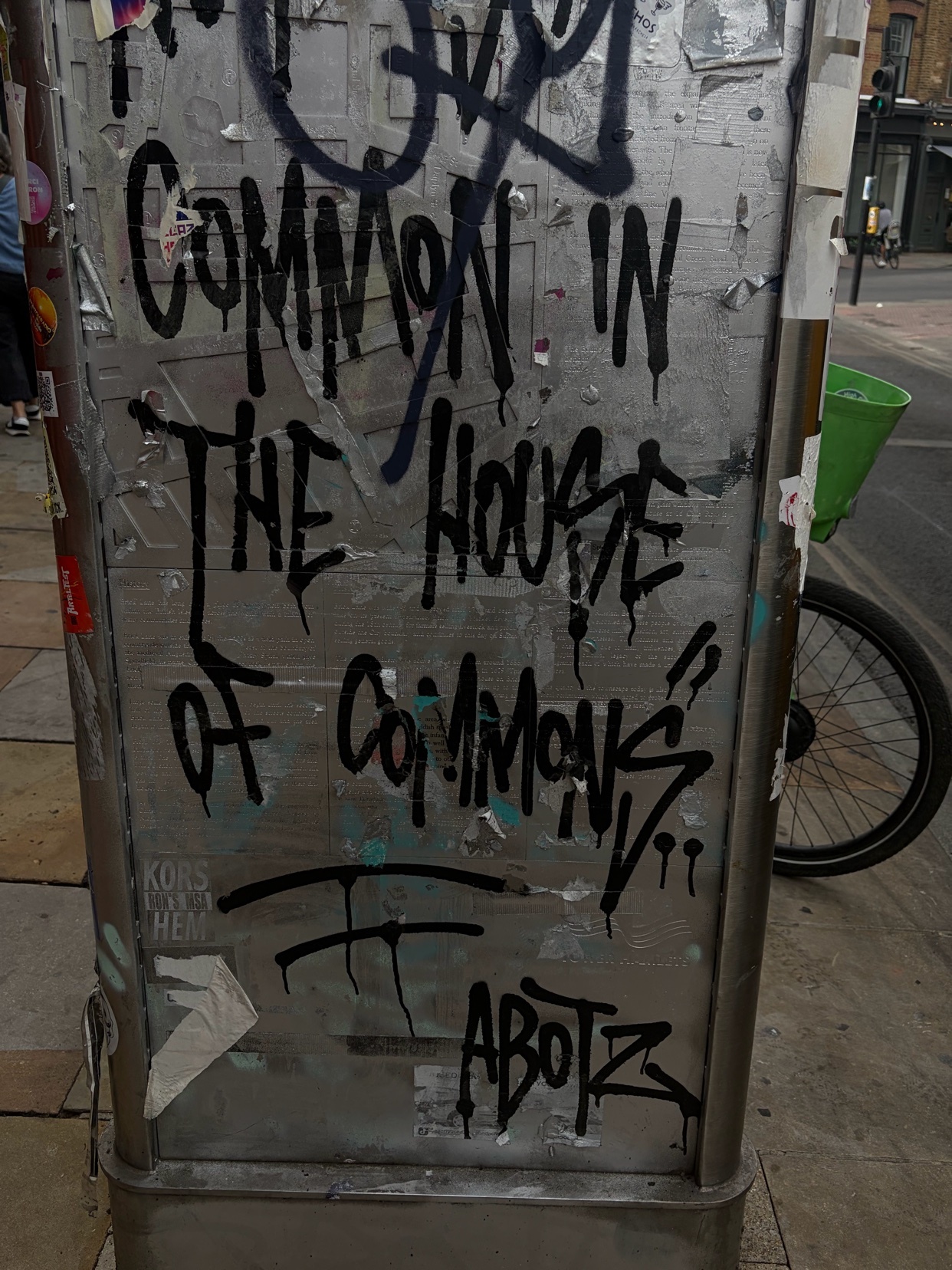 Graffiti art covers a metal pole on a city street. The artwork features bold, black lettering that reads "COMMON IN THE HOUSE OF COMMONS" with additional tags and scribbles.