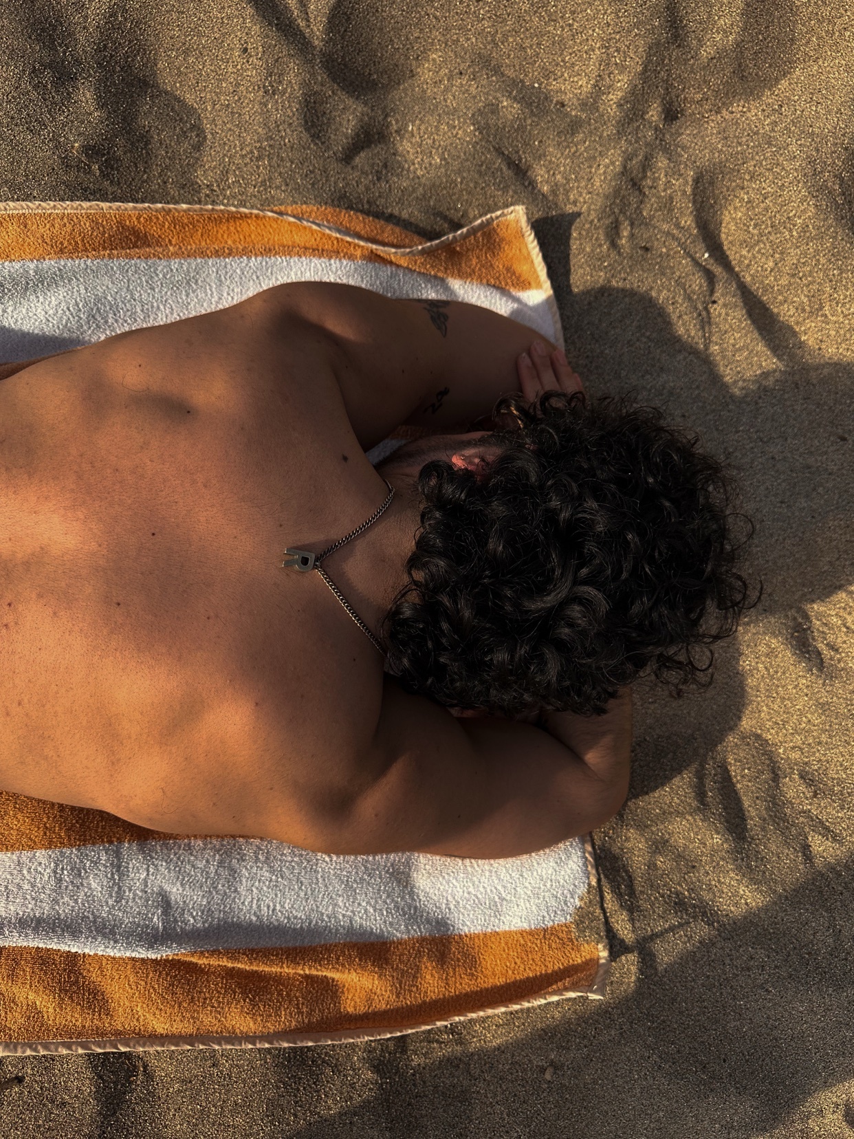 A shirtless man sunbathes on a beach towel. The photo captures his back and curly hair, with the sand and sunlight adding to the summer vibe.