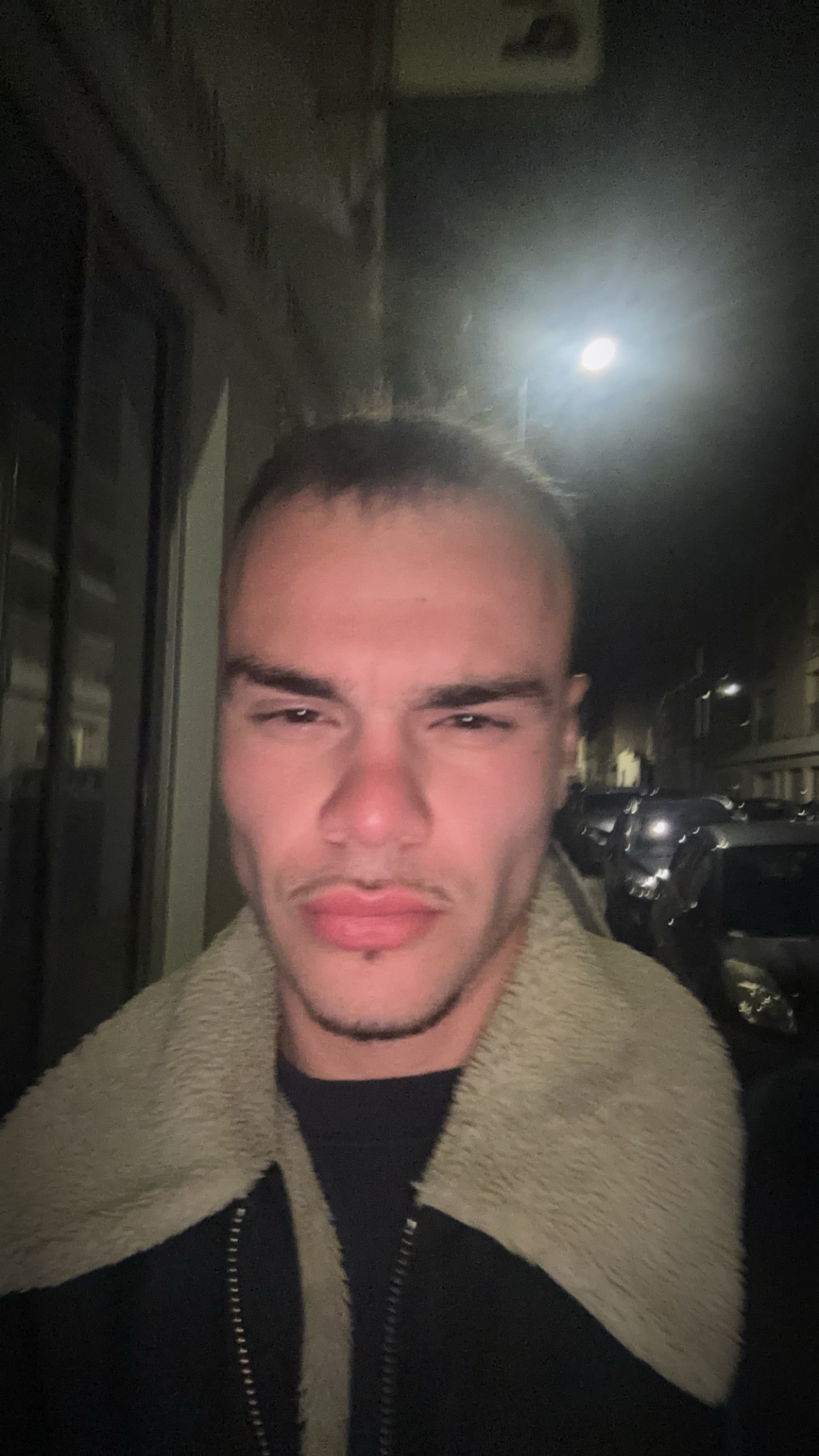 A man poses for a selfie at night. He is wearing a jacket and has dark hair and a mustache.