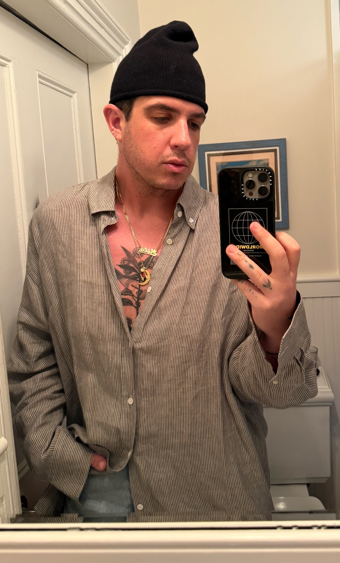 A man takes a mirror selfie in a bathroom, showcasing his style. He is shirtless, wearing a button-down shirt, a black beanie, and a gold necklace.