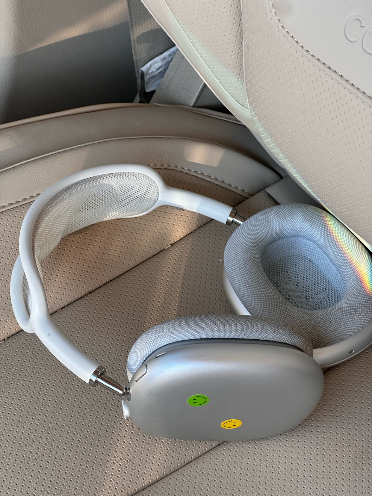 A pair of silver Apple AirPods Max headphones rests on a car seat. The headphones have white earcups and a white headband, and two stickers are visible on the right earcup: a green smiley face and a yellow smiley face.