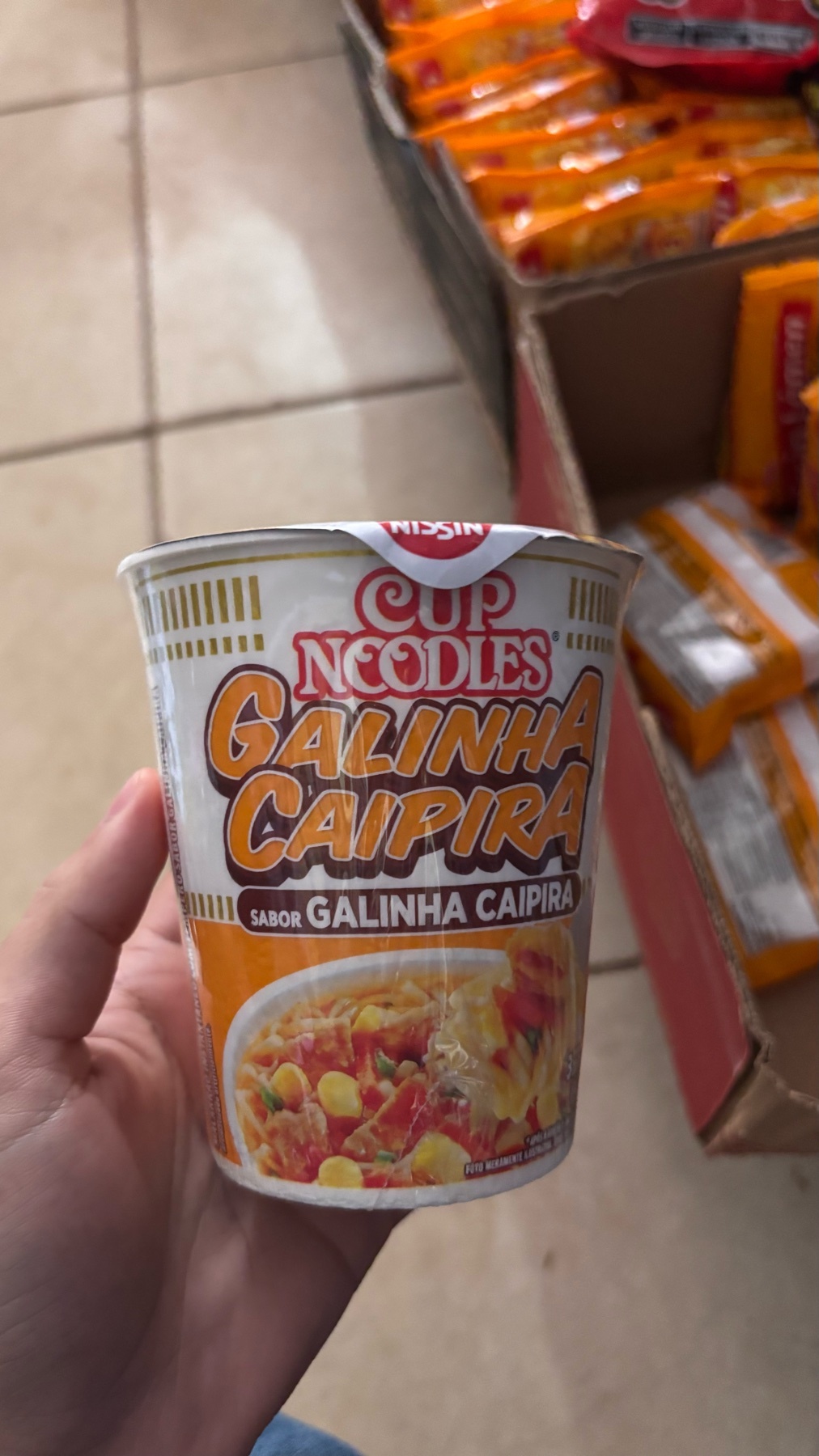 A hand holds a cup of Nissin Cup Noodles, flavor Galinha Caipira, which translates to country-style chicken. The cup is white with orange and yellow lettering and features an image of the chicken noodle soup.