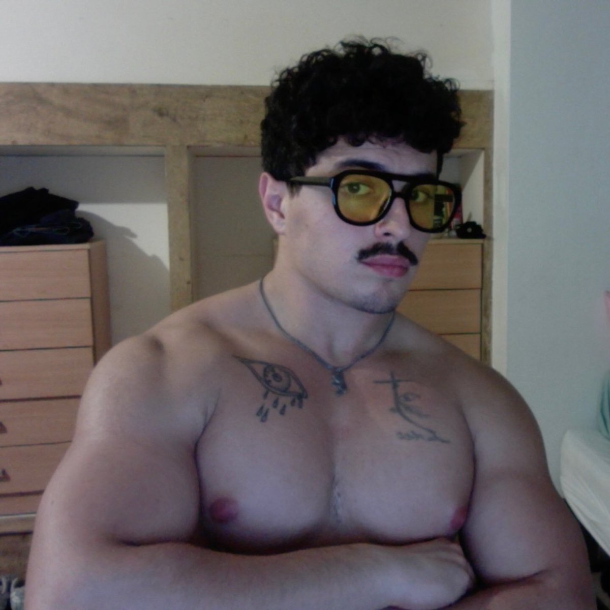 A shirtless man with muscular arms and chest poses for a selfie. He has dark curly hair, a mustache, and is wearing glasses.