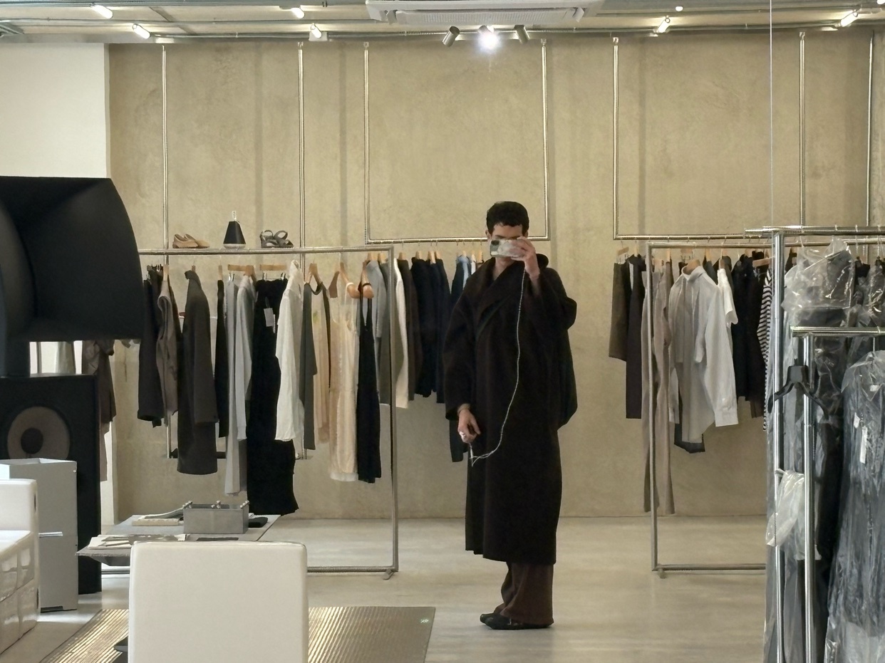 A person takes a mirror selfie in a minimalist clothing boutique. Racks of neutral-toned apparel are visible in the background.