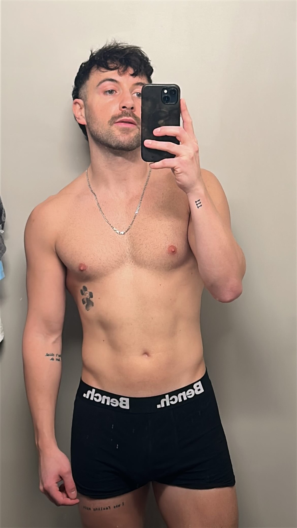 A man takes a mirror selfie, shirtless and wearing black underwear. He has dark hair, a mustache, and several tattoos.