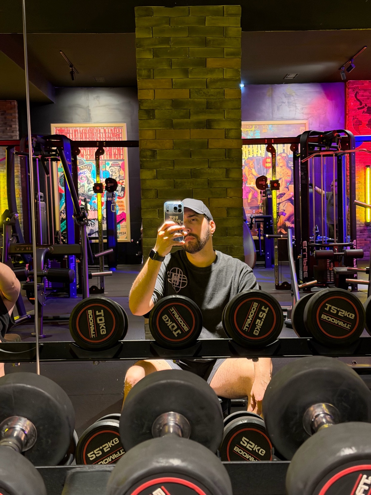 A man in a baseball cap and t-shirt takes a selfie in a gym, with rows of dumbbells in the foreground and weight machines in the background.