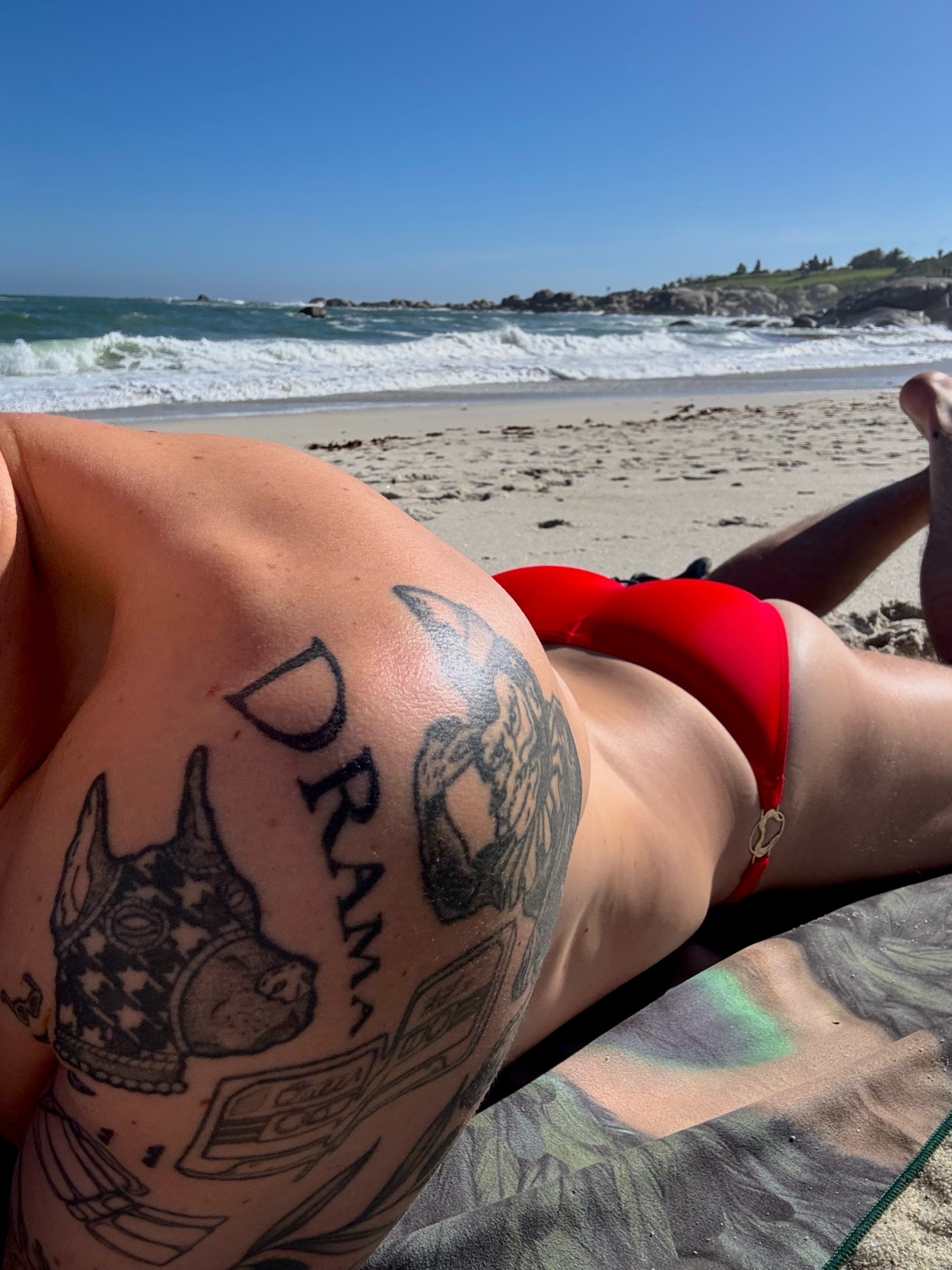 A woman in a red bikini lies on a beach towel, facing away from the camera. Her back and shoulder are visible, adorned with tattoos of dogs and the word "DRAMA". The ocean and a rocky coastline are in the background under a clear blue sky.