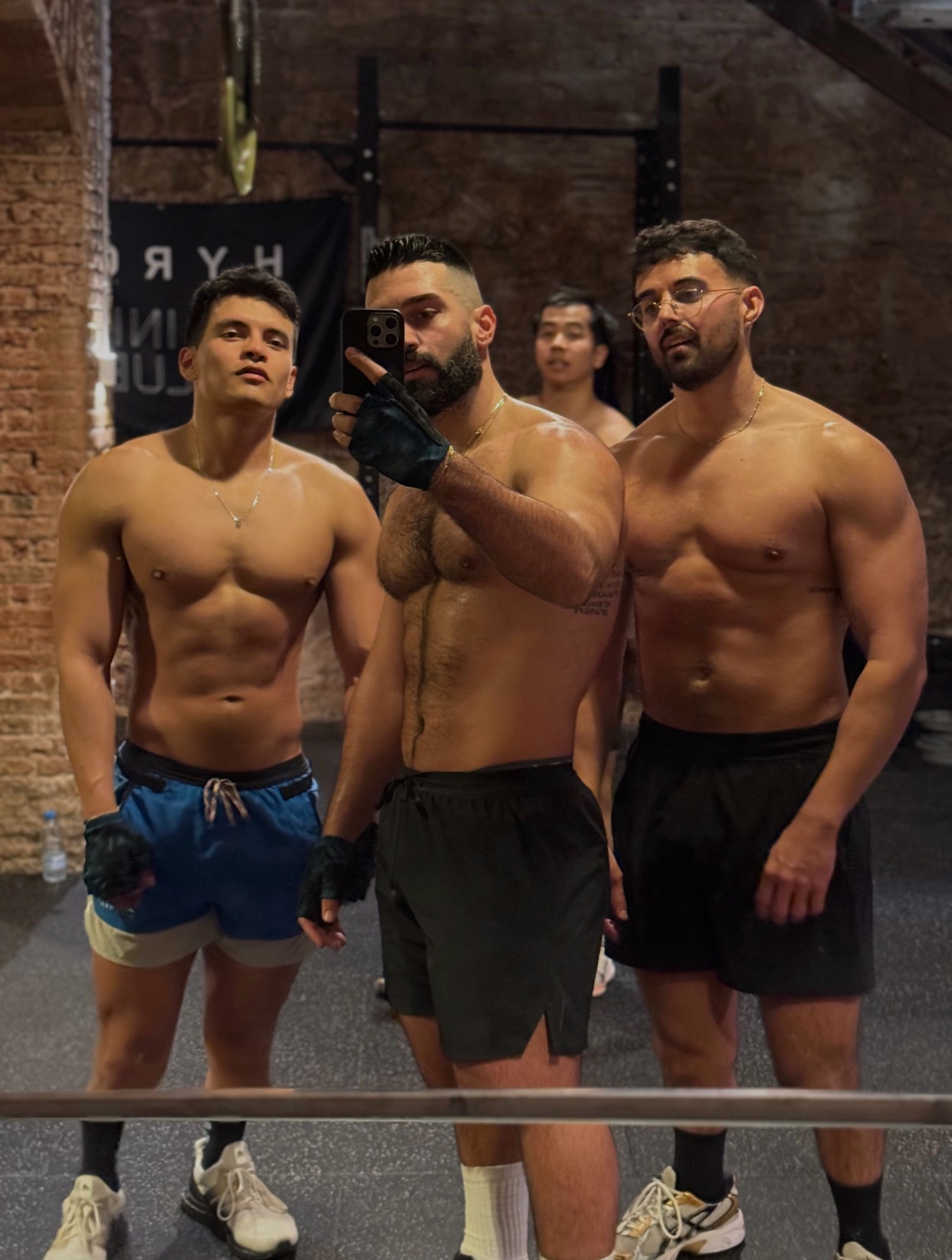Three muscular men pose for a selfie in a gym. They are all shirtless and wearing athletic shorts, with one man in the foreground holding the phone and wearing boxing gloves.