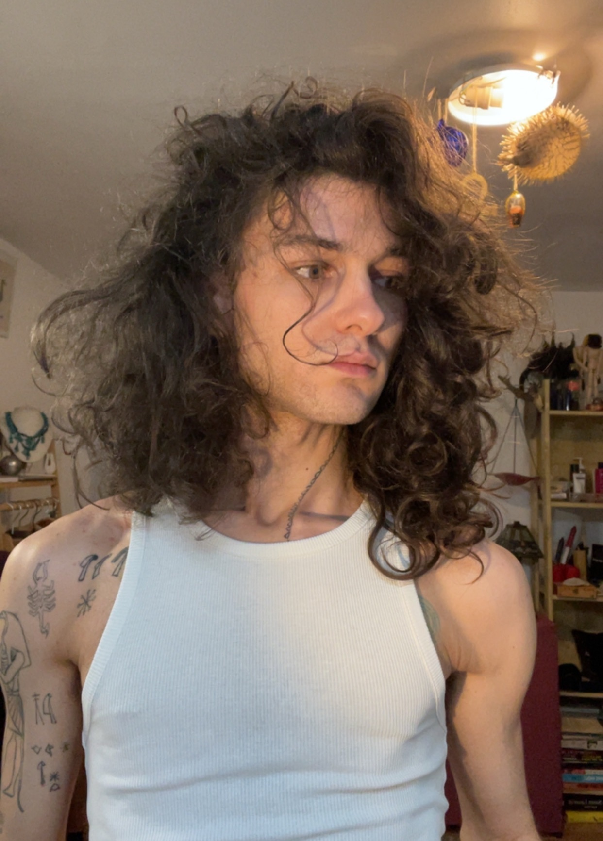 A man with long, curly hair poses shirtless in a tank top. He has tattoos on his arm.