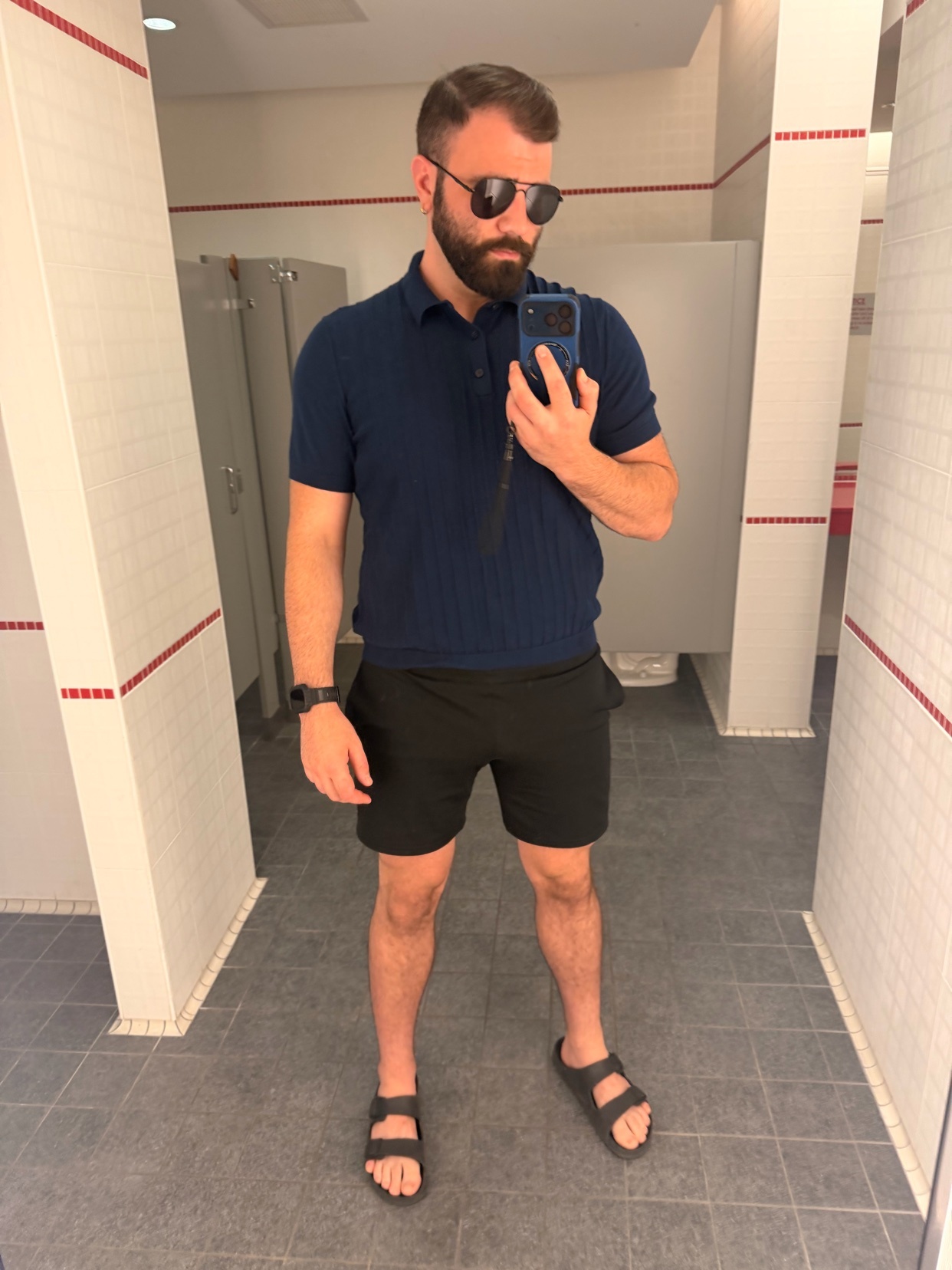 A man with a beard and sunglasses takes a mirror selfie in a locker room. He is wearing a navy blue polo shirt, black shorts, and sandals.