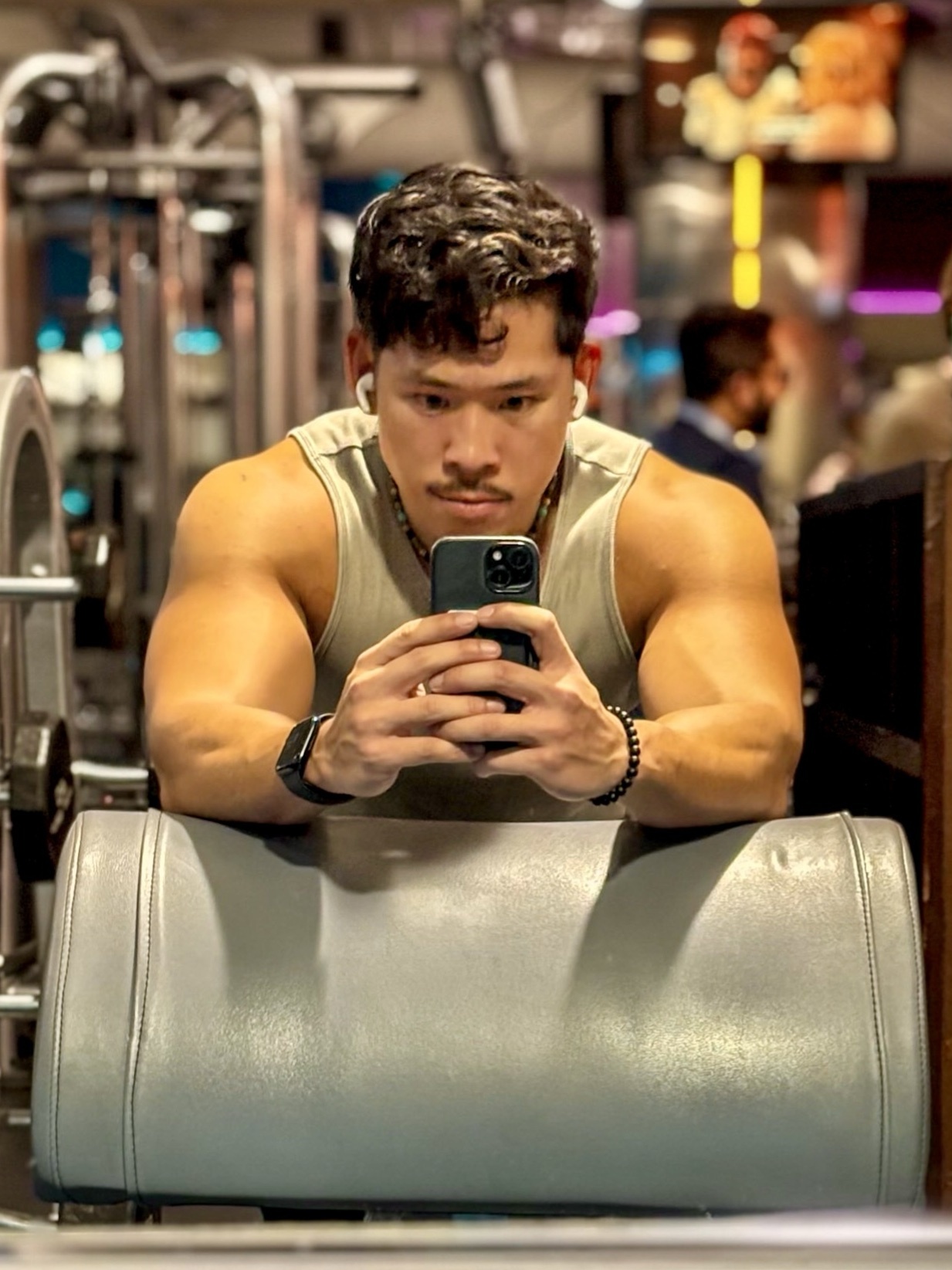 A man takes a selfie at the gym, showcasing his muscular physique. He is shirtless and flexing his biceps.