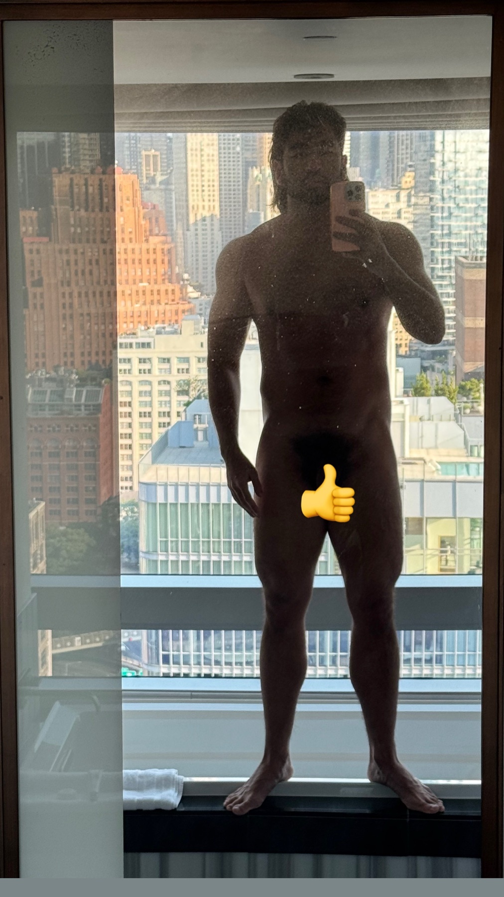 A muscular man takes a mirror selfie in front of a cityscape. He is shirtless and has a thumbs-up emoji covering his lower body.