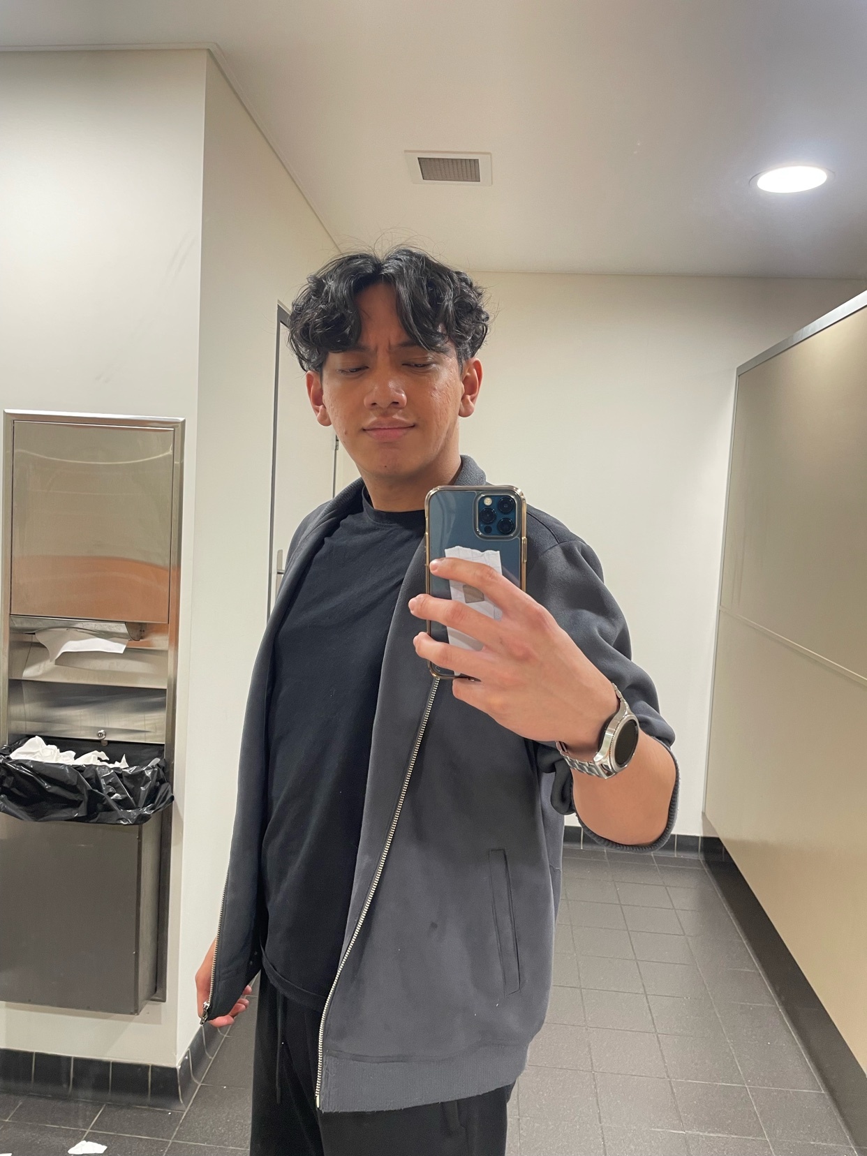 A man takes a selfie in a bathroom. He is wearing a black t-shirt and a gray jacket.