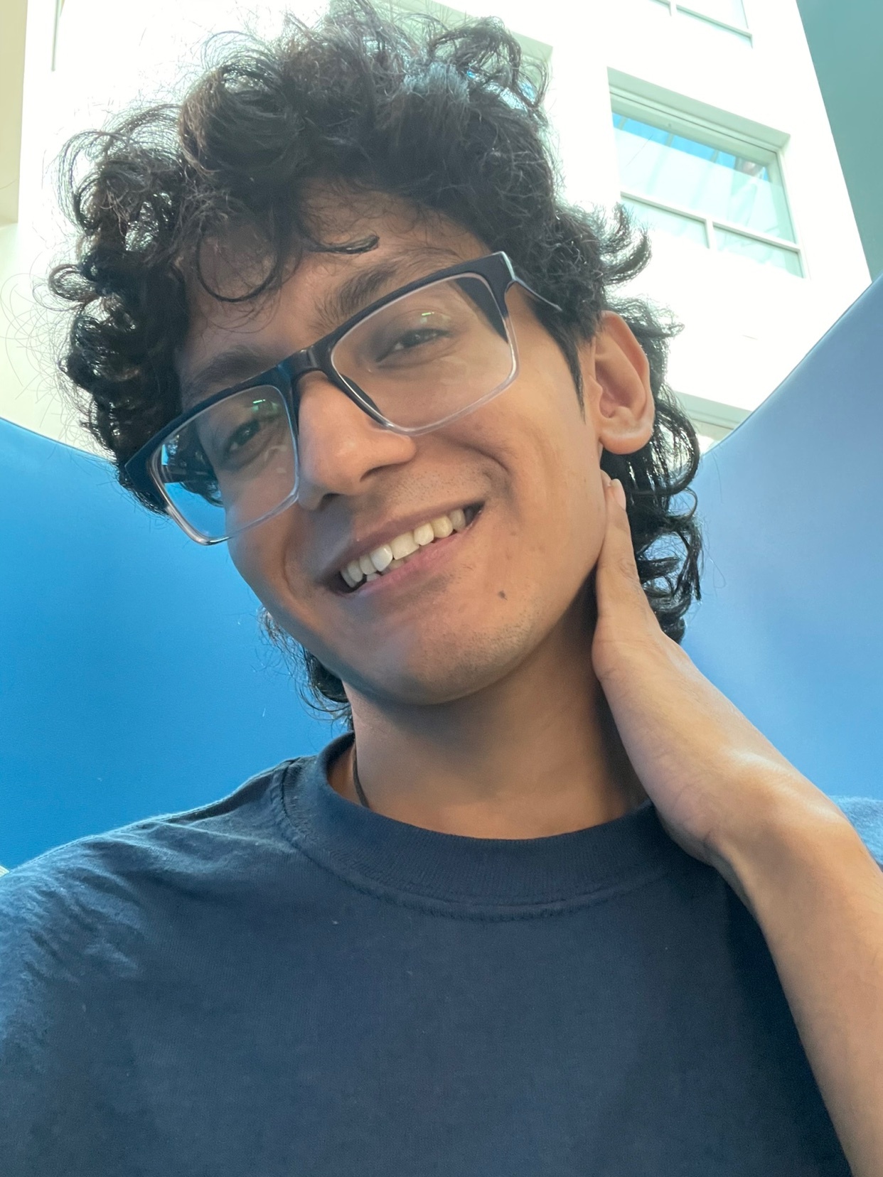 A close-up selfie of a young man with curly hair and glasses, smiling at the camera. He is wearing a blue shirt.