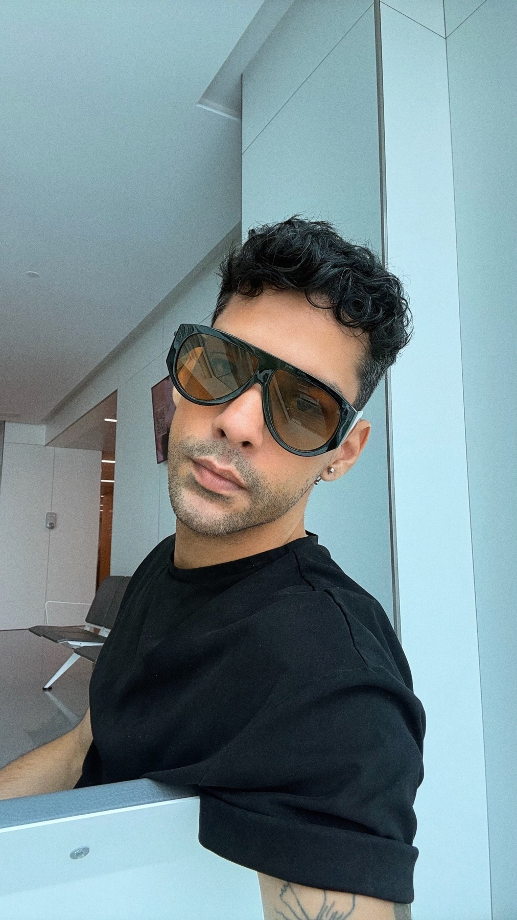A man with curly dark hair and sunglasses poses for a selfie in a modern, brightly lit interior space. He is wearing a black t-shirt and has a tattoo visible on his arm.