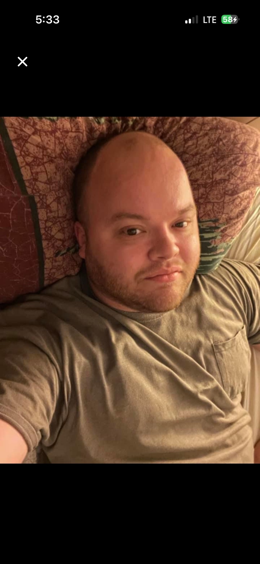 A man with a bald head and a beard lies on a patterned pillow, looking directly at the camera. He is wearing a plain t-shirt.