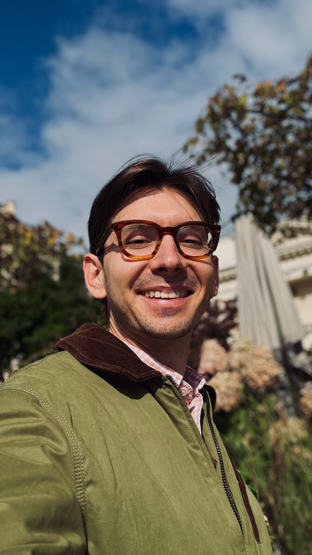 A man with dark hair and glasses smiles at the camera outdoors on a sunny day. He is wearing a green jacket over a collared shirt.