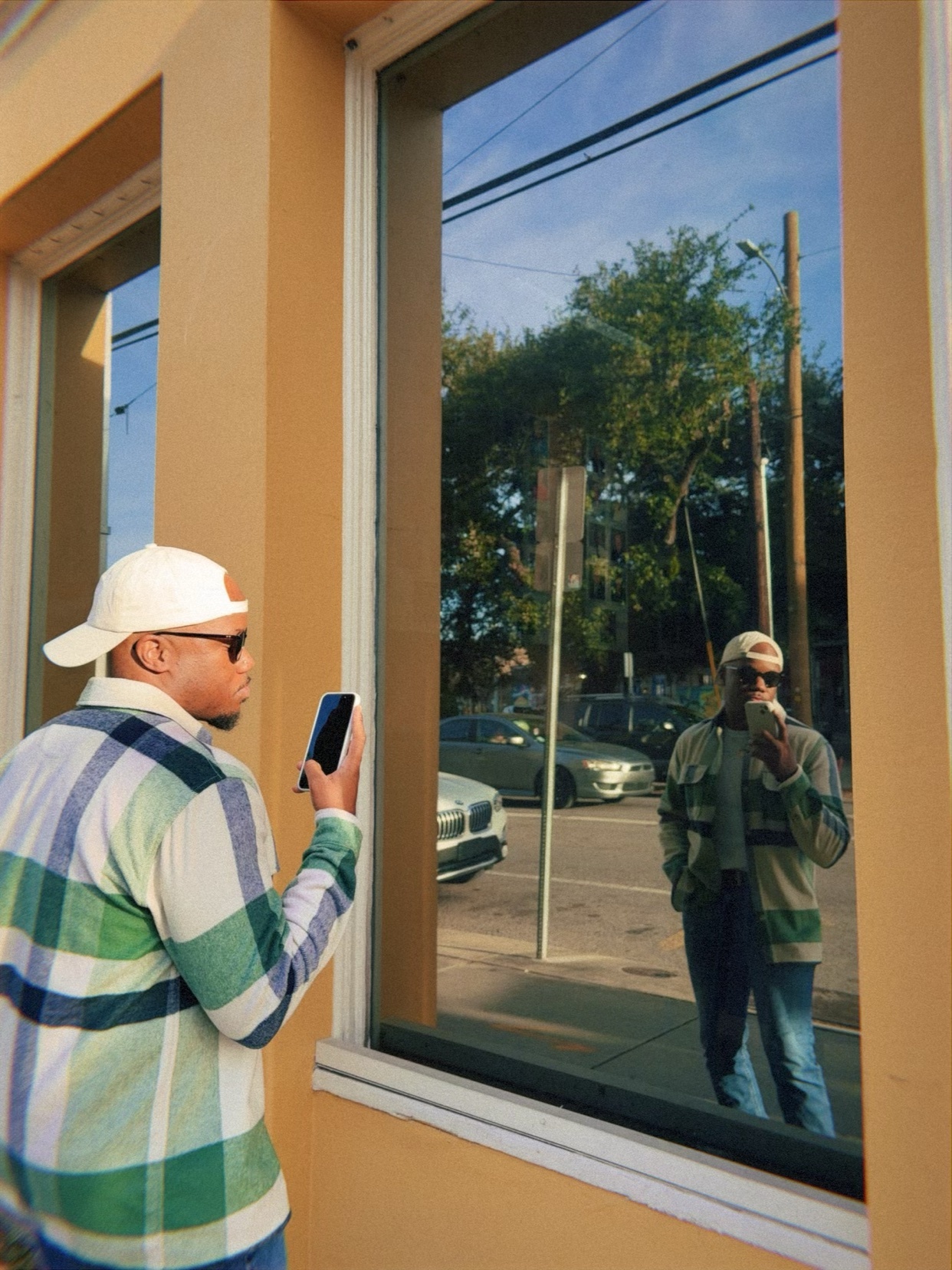 A man takes a selfie in front of a window, capturing his reflection. He's wearing a plaid shirt, cap, and sunglasses.