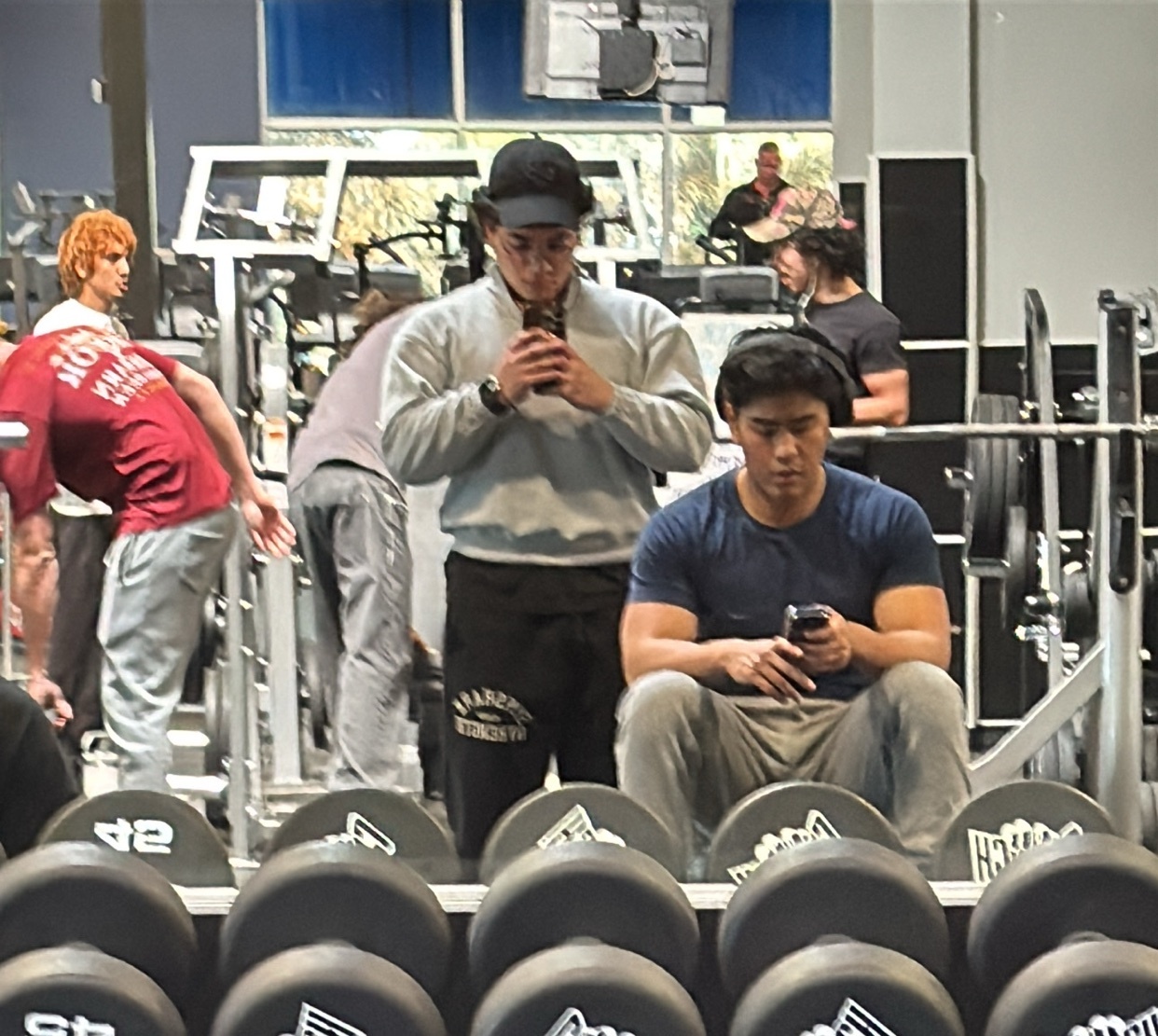 A group of men are in a gym, some working out and others taking selfies. The photo is taken in front of a mirror, showing the gym equipment.