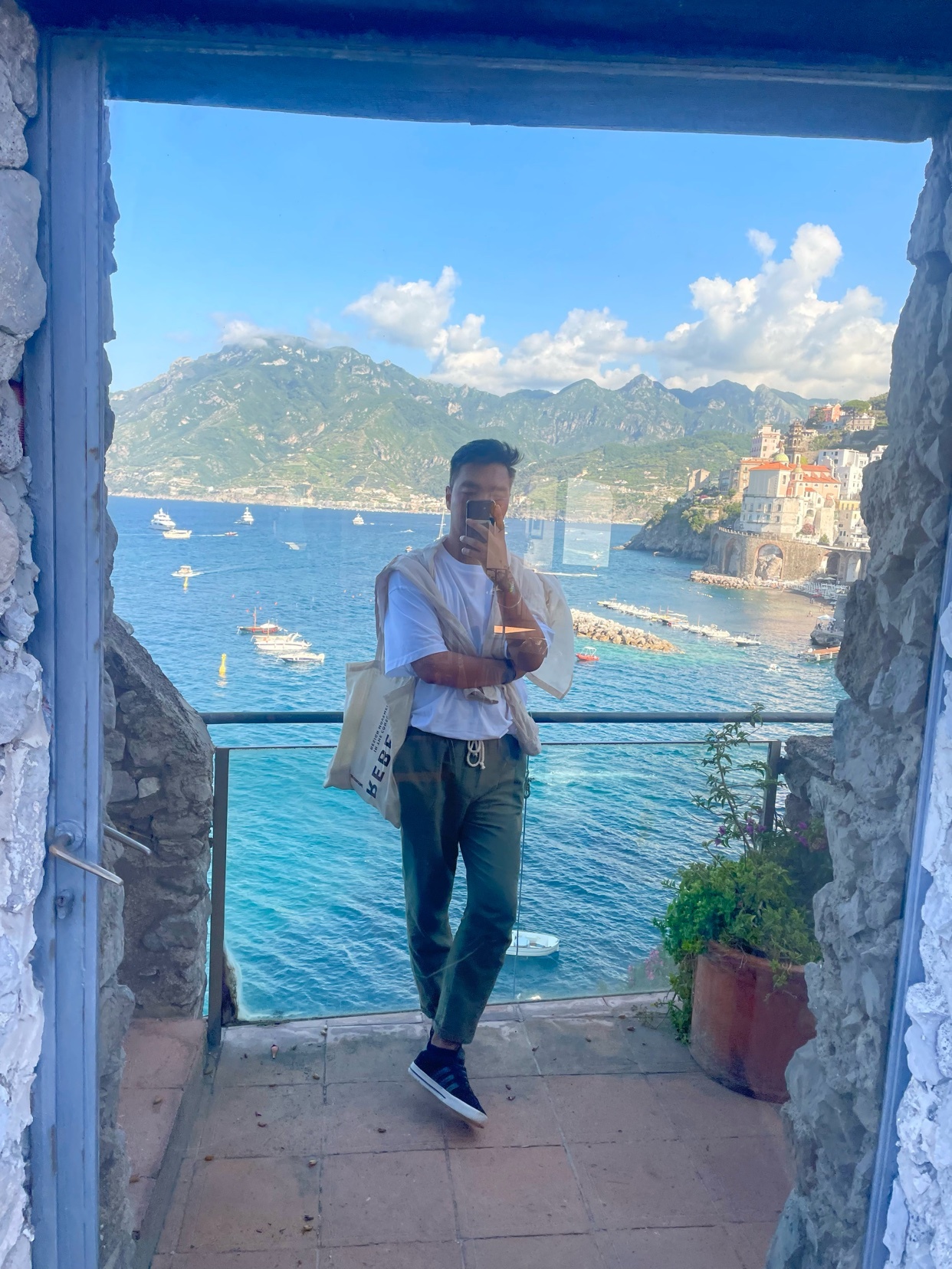 A man poses for a selfie in front of a stunning ocean view with boats and mountains in the background. He is wearing casual clothing and a tote bag is slung over his shoulder.