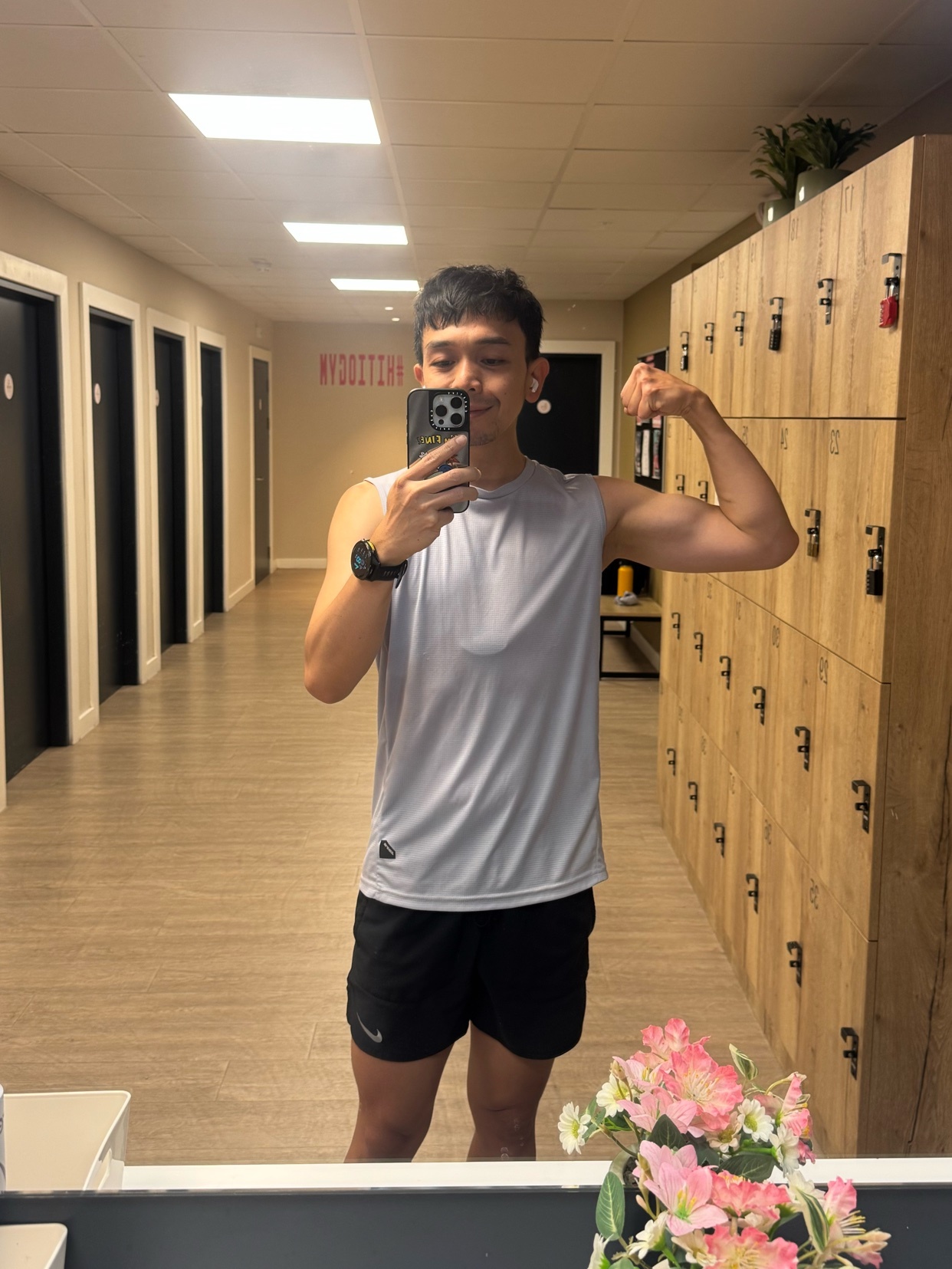 A man takes a mirror selfie in a gym locker room, flexing his bicep. He is wearing a grey tank top and black shorts.