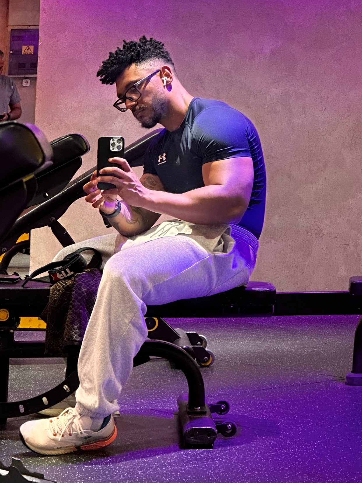 A man with glasses and a beard takes a selfie in a gym while sitting on a weight bench. He is wearing a dark t-shirt and grey sweatpants, with his muscular arms visible.