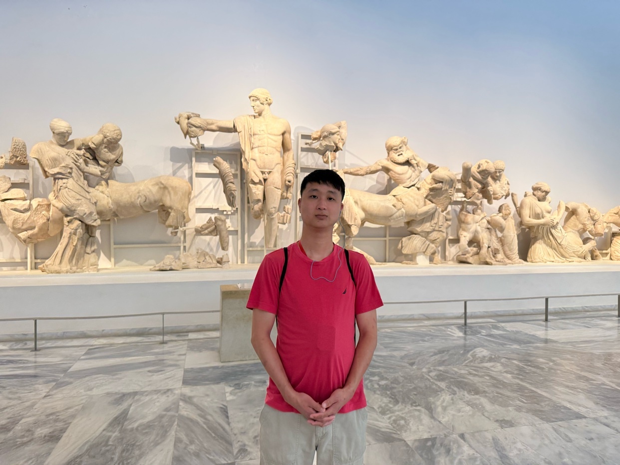 A young man stands in front of a display of ancient Greek sculptures in a museum. The sculptures depict mythological scenes and figures, carved from marble.
