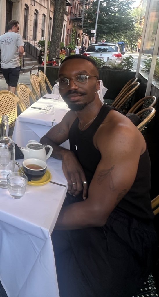 A man sits at a table outside a cafe, looking at the camera. He wears glasses and a black tank top.