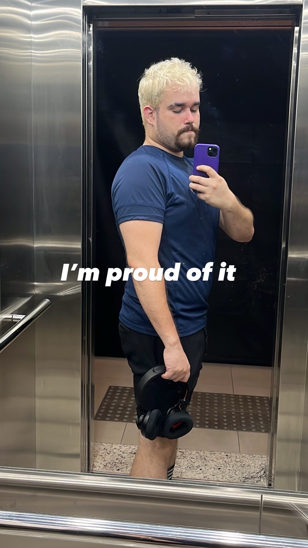 A man with blond hair and a beard takes a selfie in an elevator, holding headphones and a weight plate. He is wearing gym clothes and appears proud of his progress.