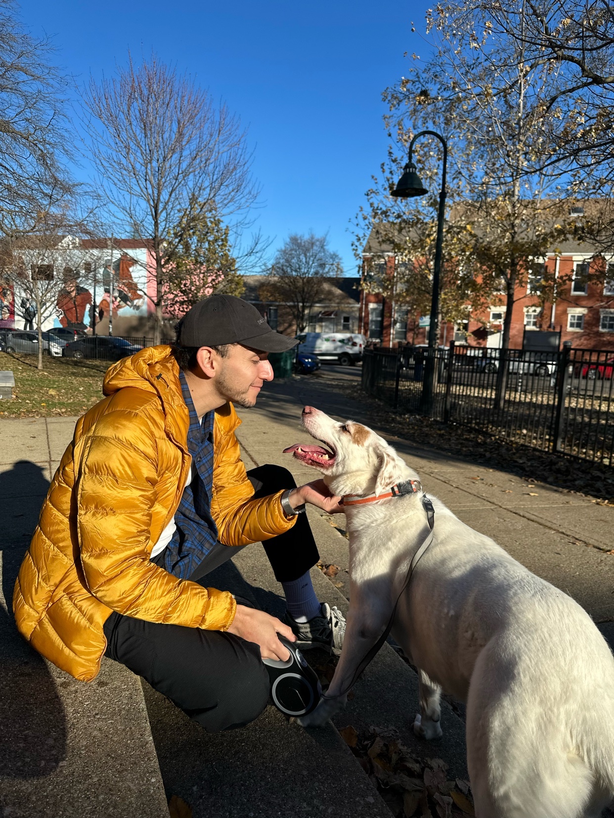 A man kneels to pet a dog in a park. The man is wearing a yellow jacket and a cap.