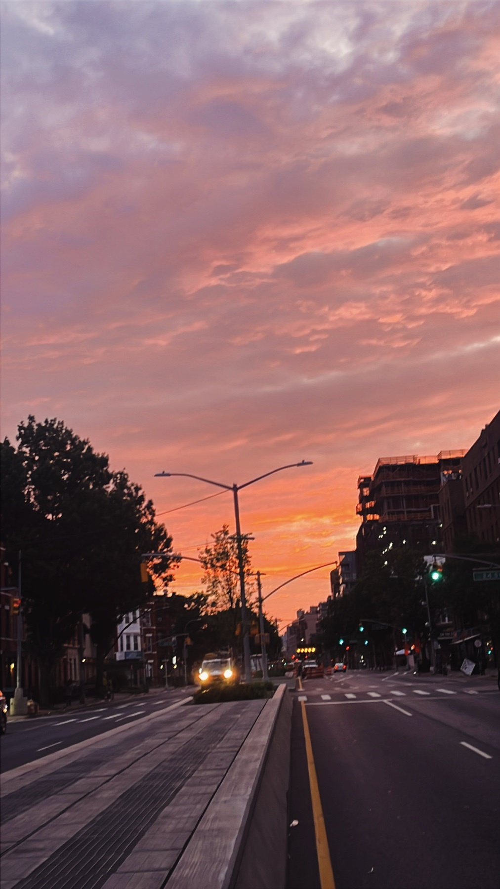A vibrant sunset paints the sky in hues of pink and orange over a city street in Brooklyn. The warm glow reflects off the clouds and illuminates the buildings and trees lining the road.